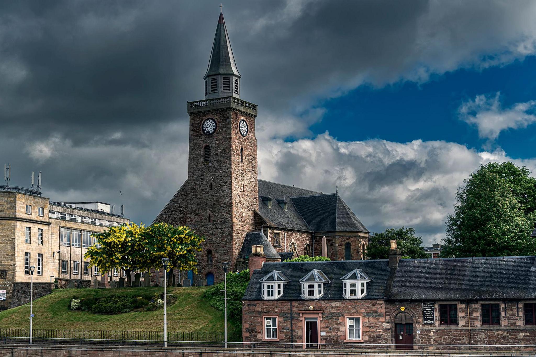 A view of Inverness' Old High Church from across the River Ness. The church is a stone-built kirk with a steeple with clock faces near the top. There is a row of traditional cottages on the street below.