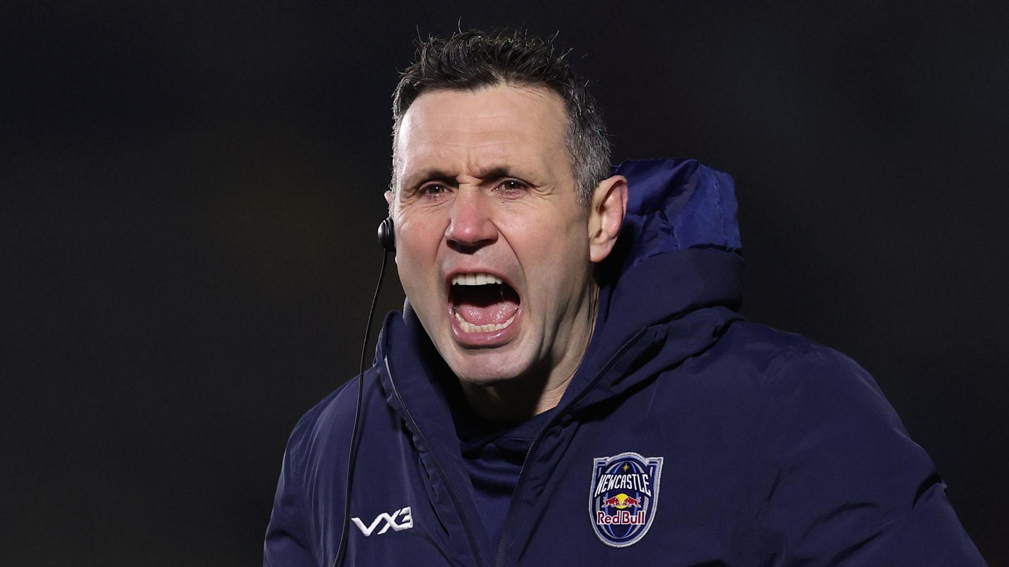 Stephen Jones, with an earpiece in his right ear and wearing a big branded navy Newcastle Red Bulls jacket, shouting instructions at his players