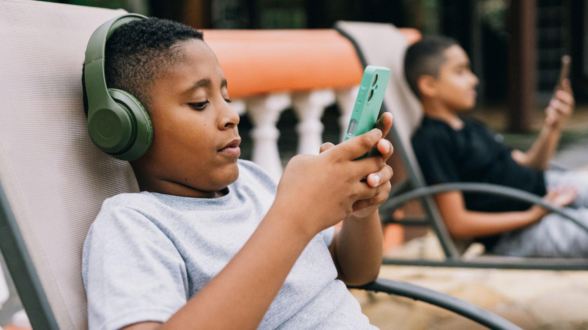 Two young boys sit on sun loungers looking at phones. The boy in the forefront wears a grey t-shirt and green headphones.