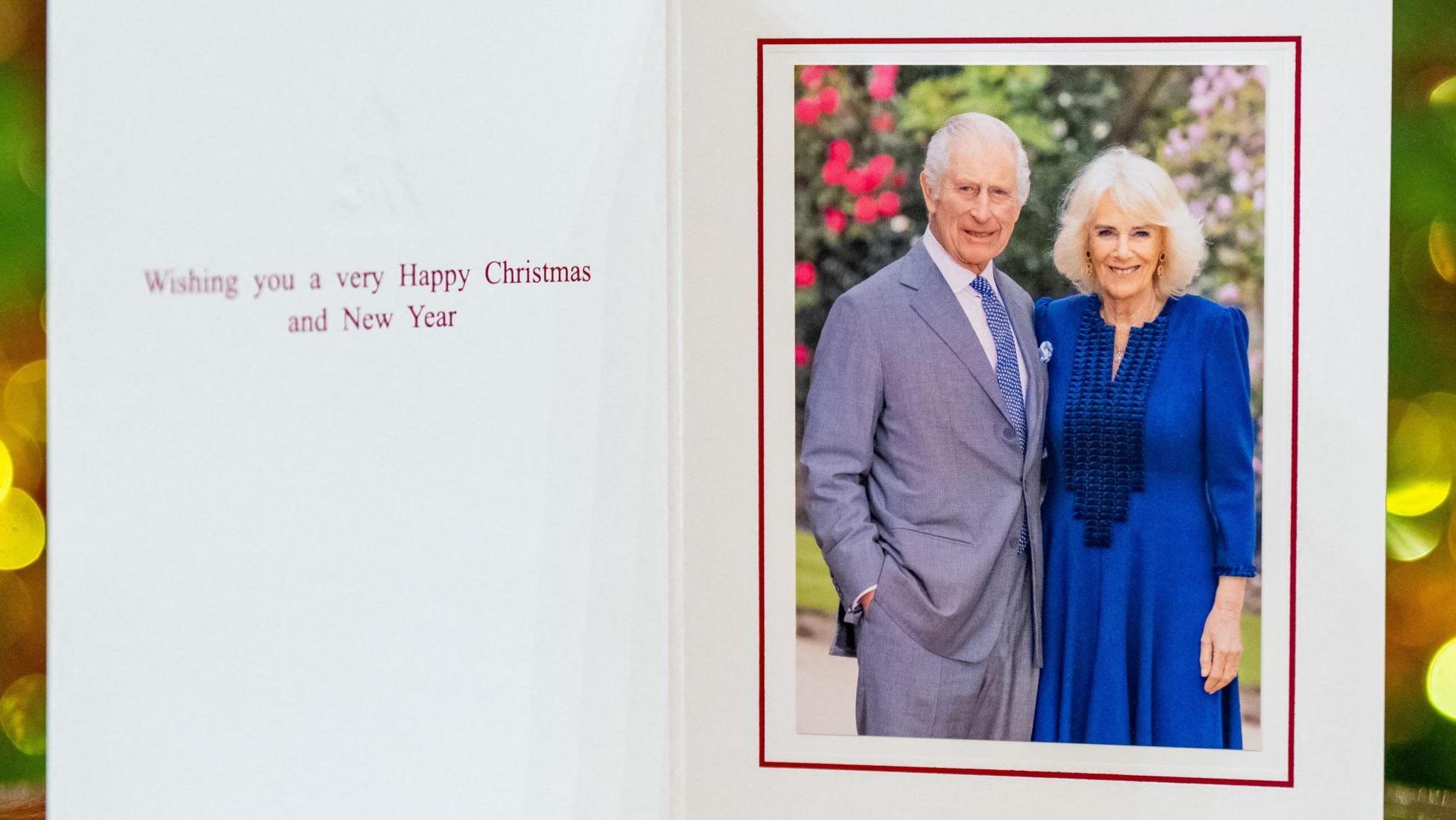 King Charles, in a grey suit, and Queen Camilla, wearing a long blue dress, are pictured in front of some shrubs in a Christmas card bearing the message: Wishing you a very Happy Christmas and New Year.