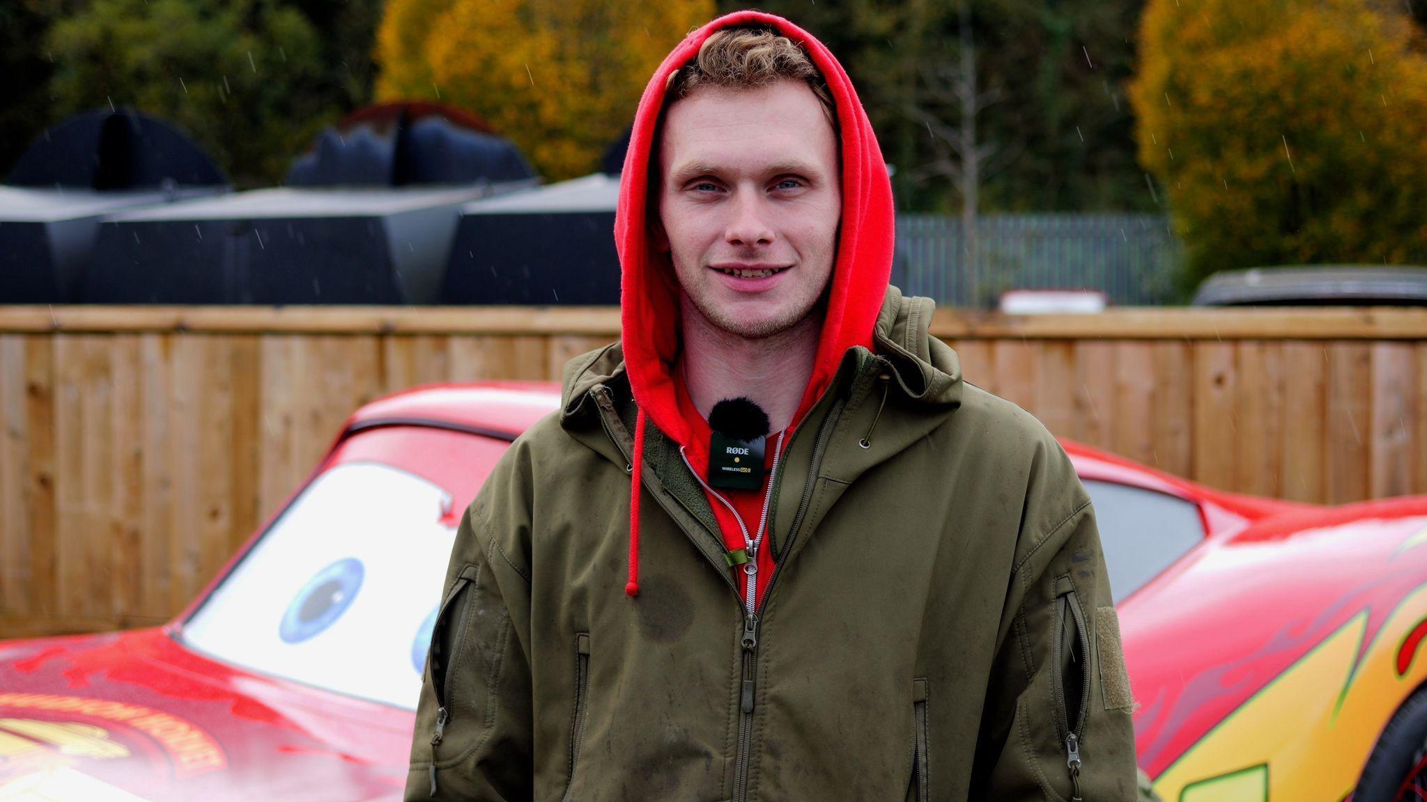 A man looking into the camera, stood in front of a bright red car. He is wearing a red hoodie and a dark green coat, and has a wireless microphone attached to his collar.