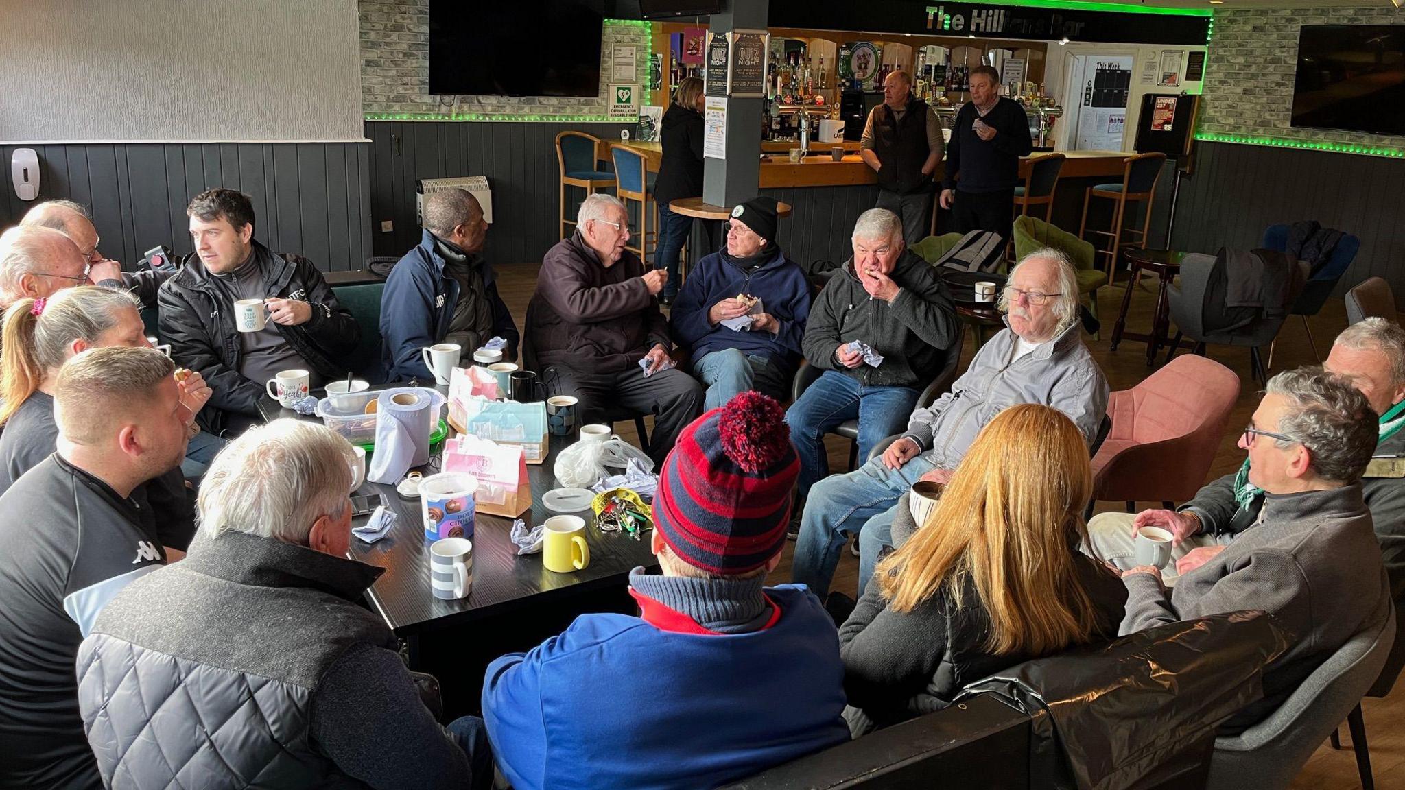 A group of around 15 people sit around a table in the lounge. They are drinking tea and chatting to one another. 