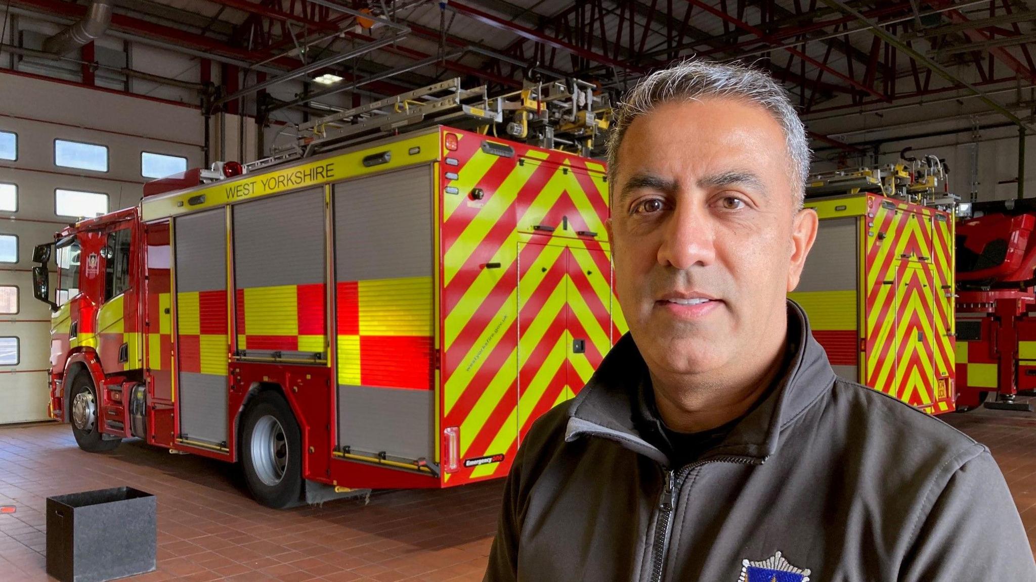 A man with silver hair wearing a grey fire service jacket with an upturned collar in a fire station with two fire engines with aluminium ladders on their roofs.  