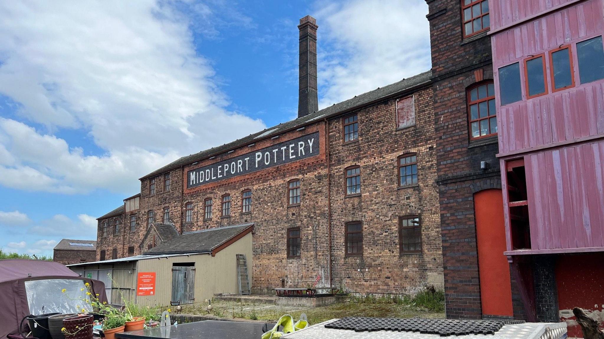 The brown brick building has a large Middleport Pottery sign in black and white high up. A chimney can be seen behind and a red painted door and a pink building can be seen on the right of the image at ground level. The building appears to be by water with a small maroon boat moored a few feet away.