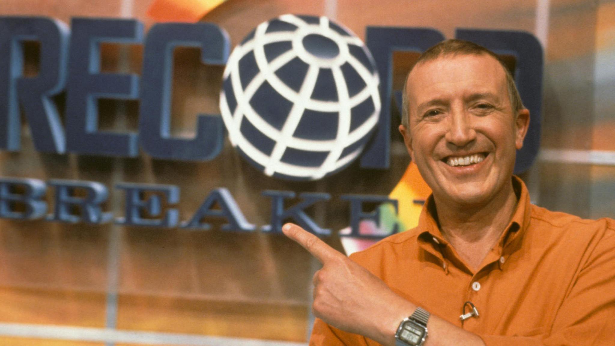 Roy Castle dressed in orange shirt with short brown hair pointing at Record Breaker logo