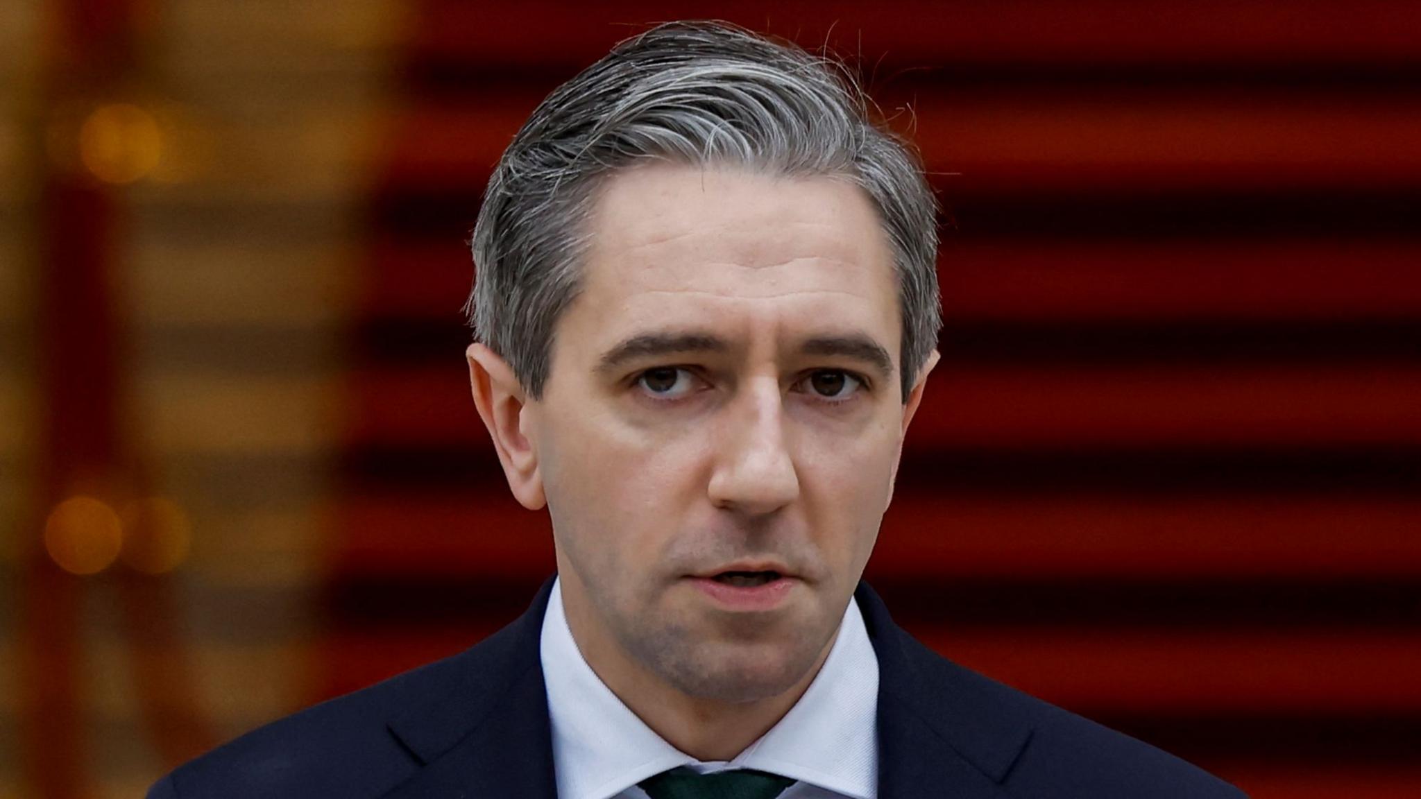 Simon Harris - a man with grey hair wearing a suit and white collared shirt stands with his mouth open in front of a blurred red staircase