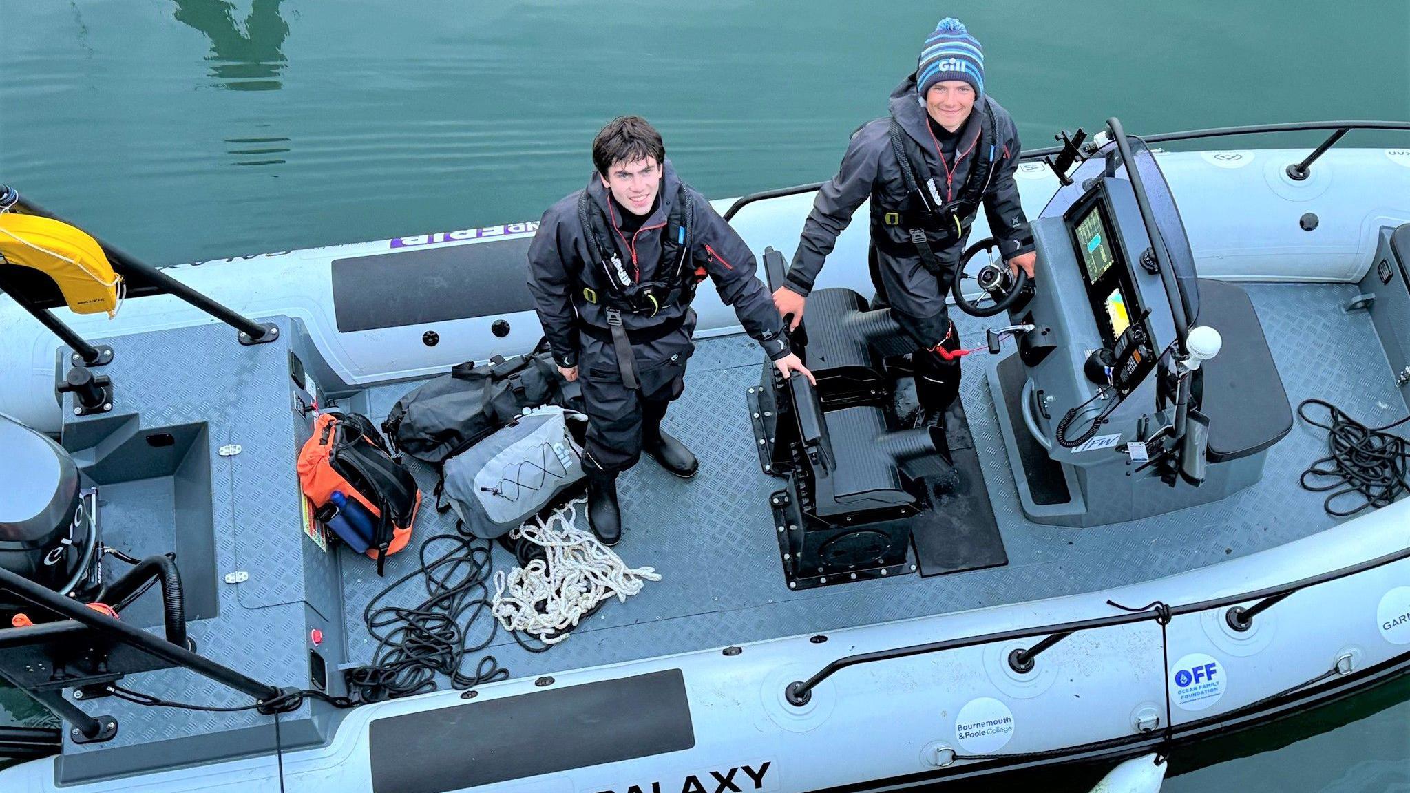 Harry Besley and his friend Charley on boat the electric boat, taken from above. You can see backpacks and coiled rope sitting in the bottom of the boat. The pair are both wearing black waterproof overalls and life jackets