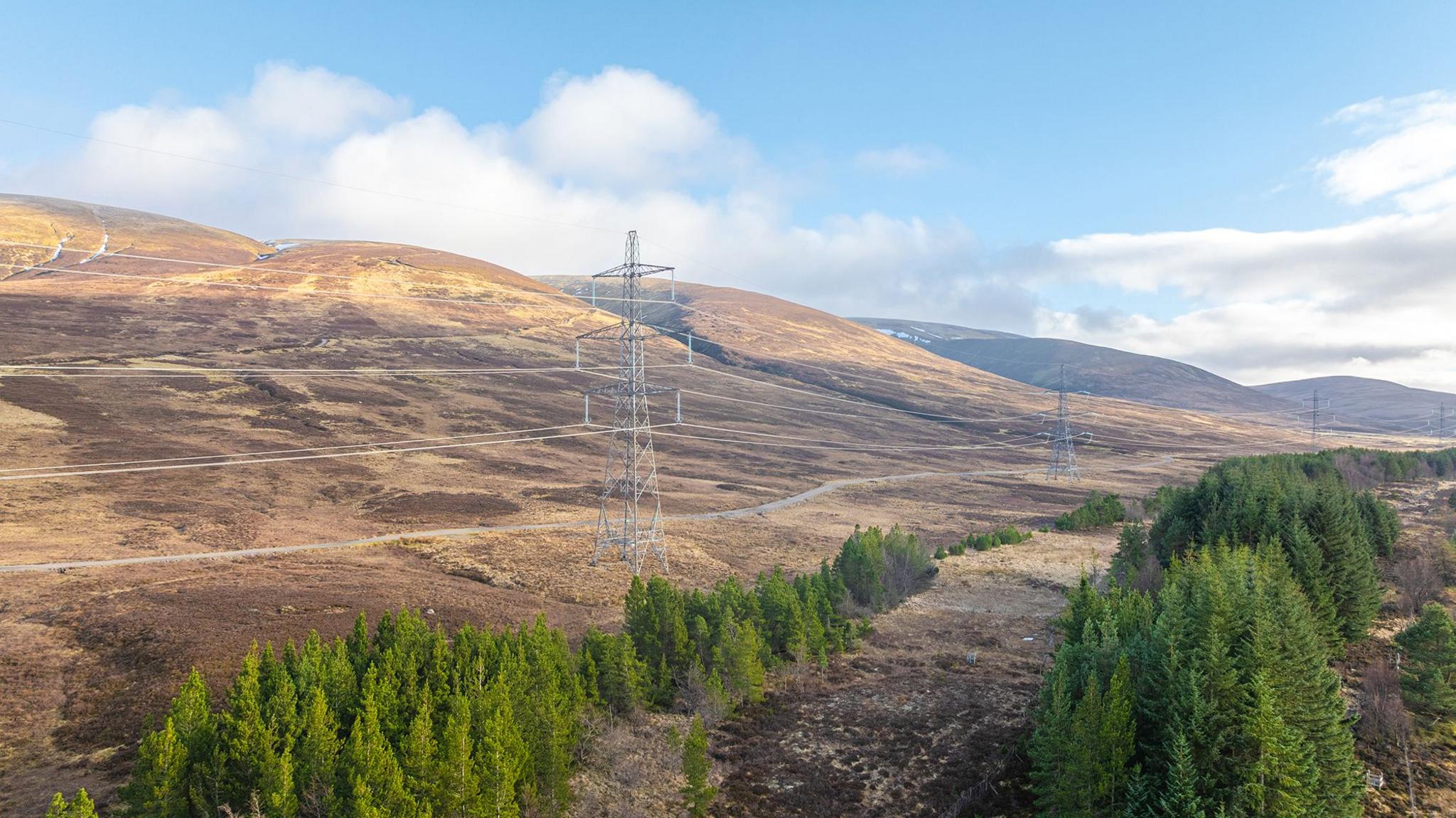 A power line and latticed pylons running through an area of trees and a hilly landscape