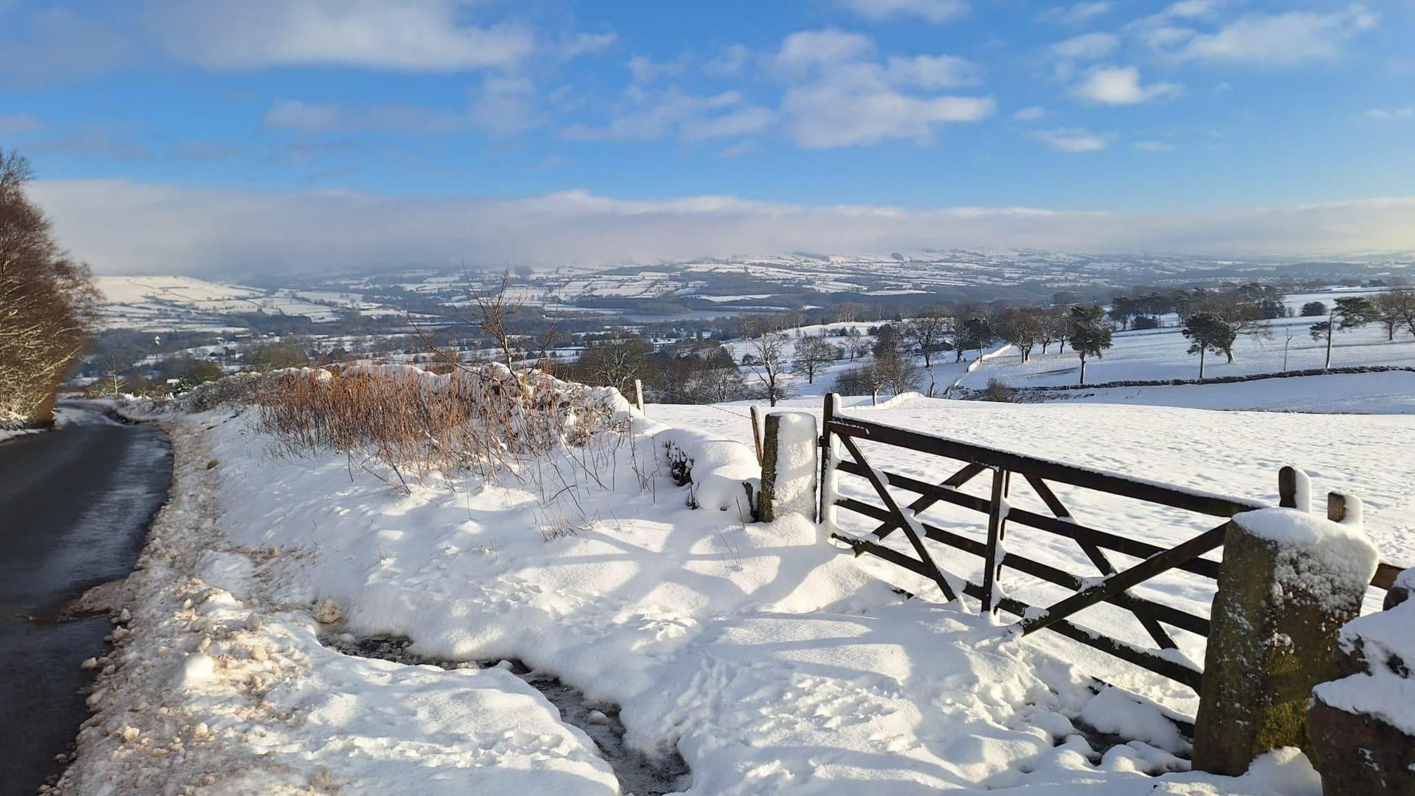 A view of snow-covered fields stretches to the horizon. A field gate is on the right with a road on the left.