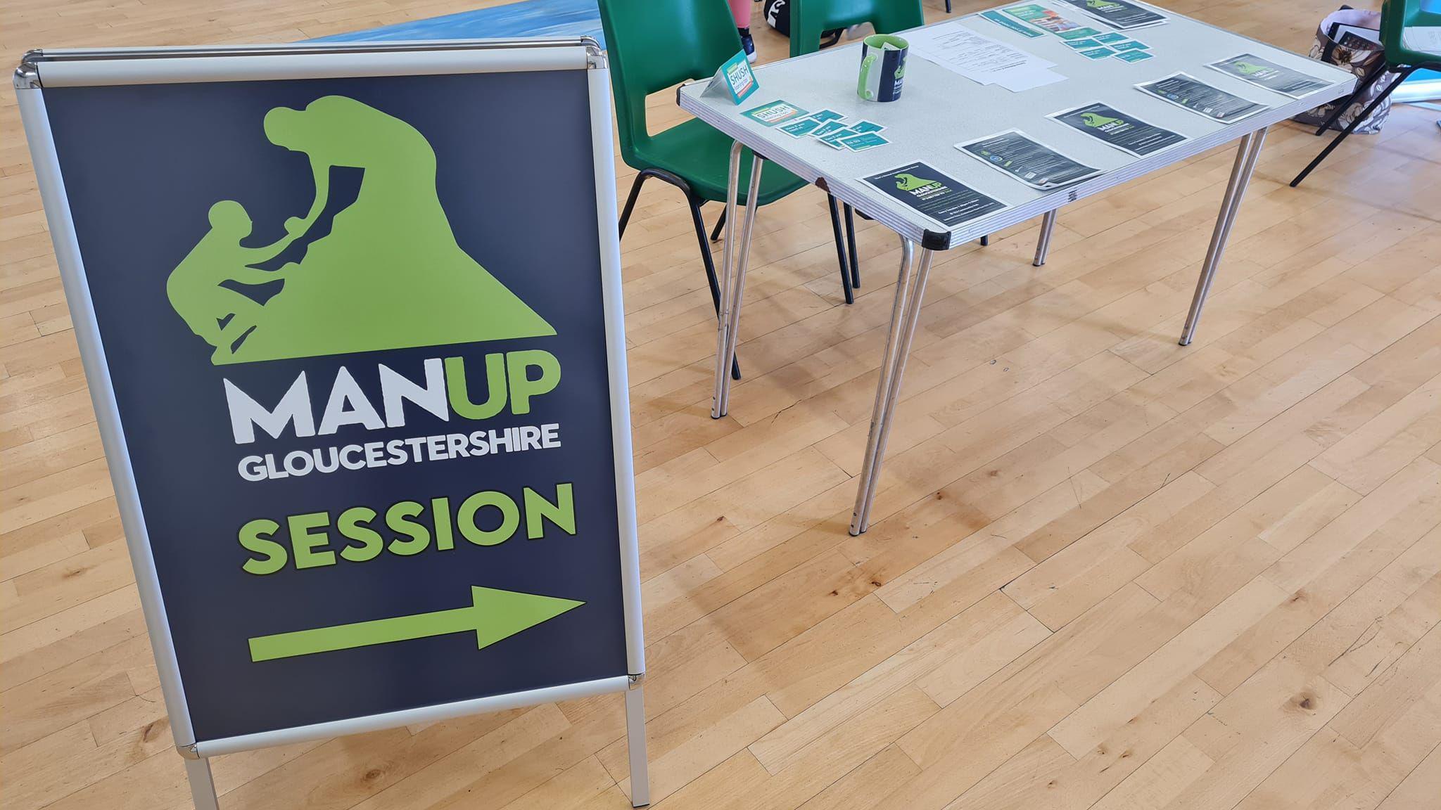 A navy blue sign which says 'MAN UP Gloucestershire session' in green and white writing. There is an arrow pointing to the right below the pictures. To the left there is a white table with leaflets placed on top of it.