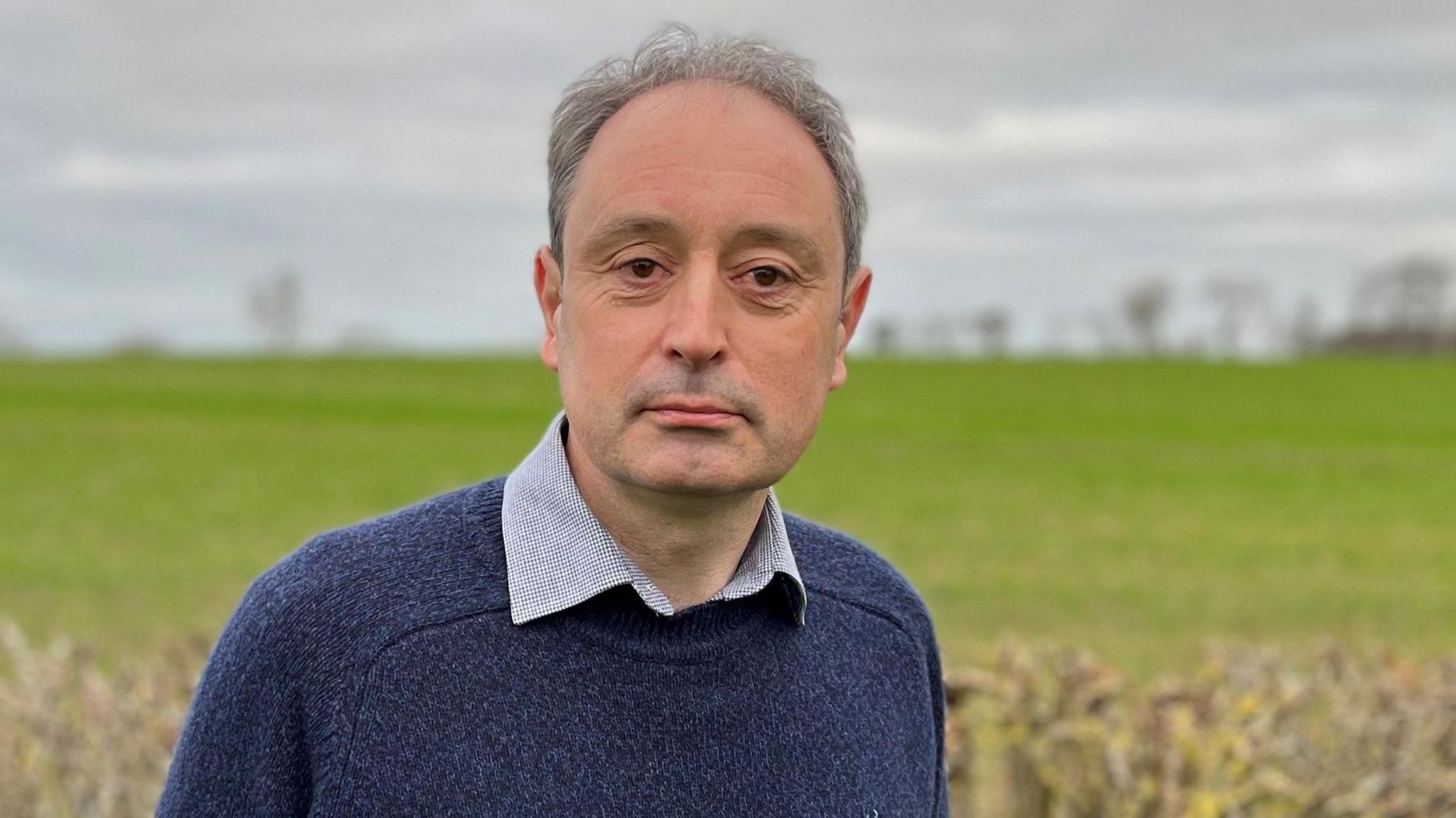 A middle-aged man with grey hair, wearing a blue jumper, standing in front of a farm field