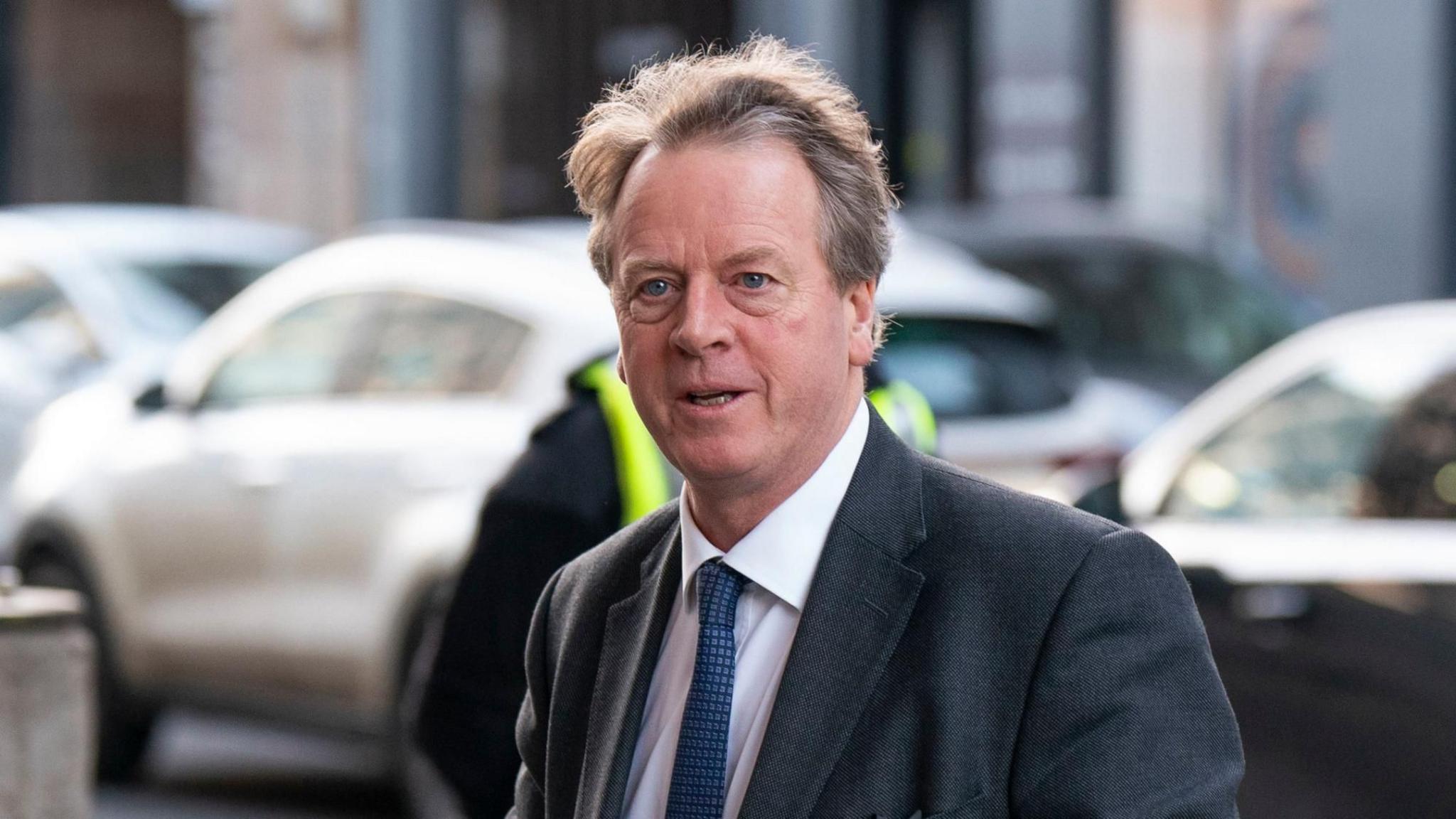 Alister Jack is wearing a dark suit jacket over a white shirt and a patterned blue tie. He has light brown/greyish hair that appears slightly tousled. The background features parked cars.