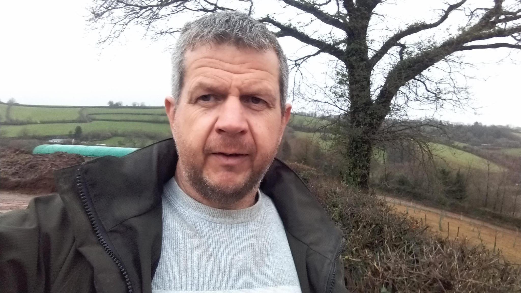 A man in a grey jumper and dark green jacket standing in front of several fields on a hillside, with a tree behind him.