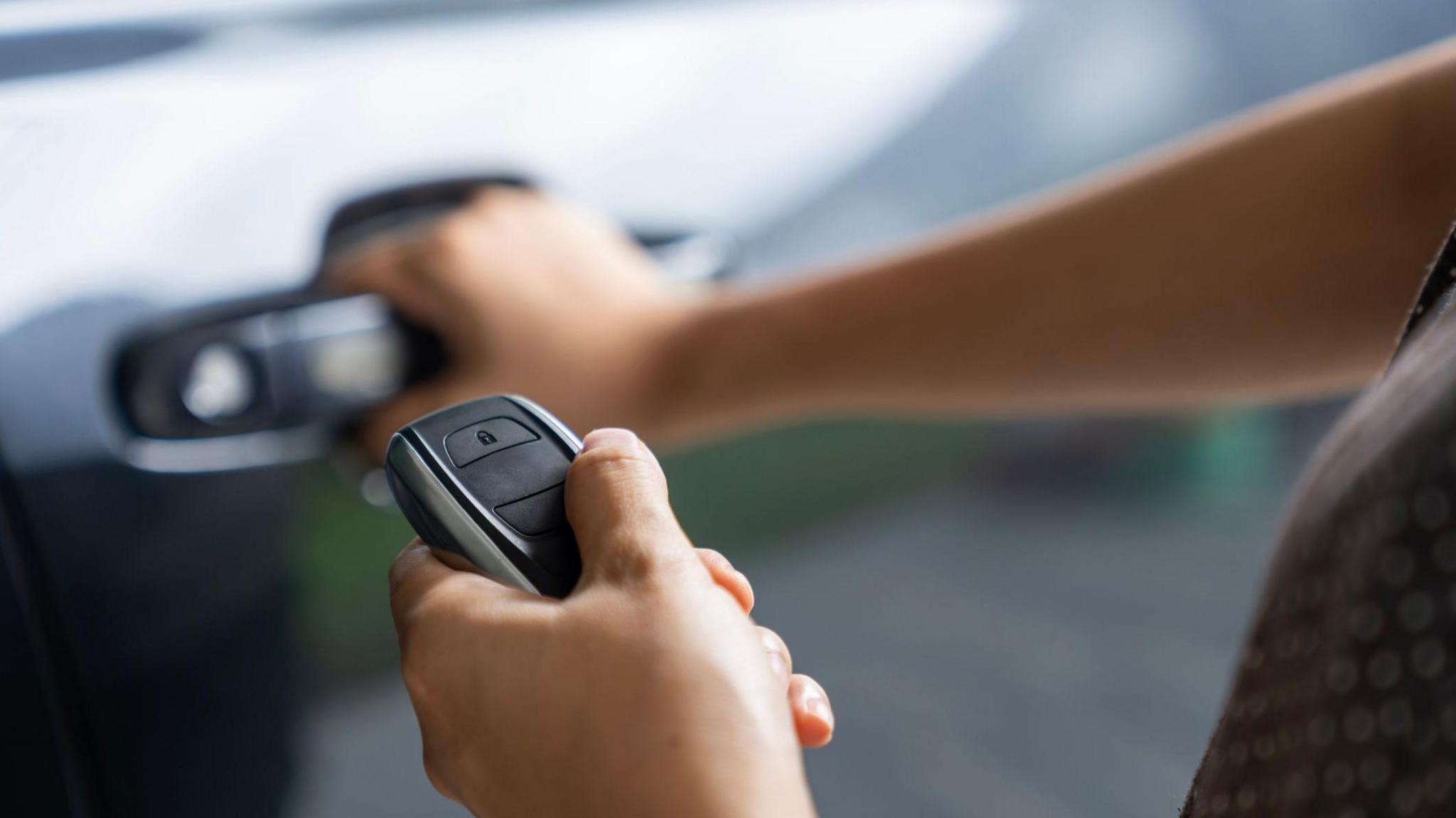 A photo of a person holding a car key with their left hand and holding the door handle of a black car. 