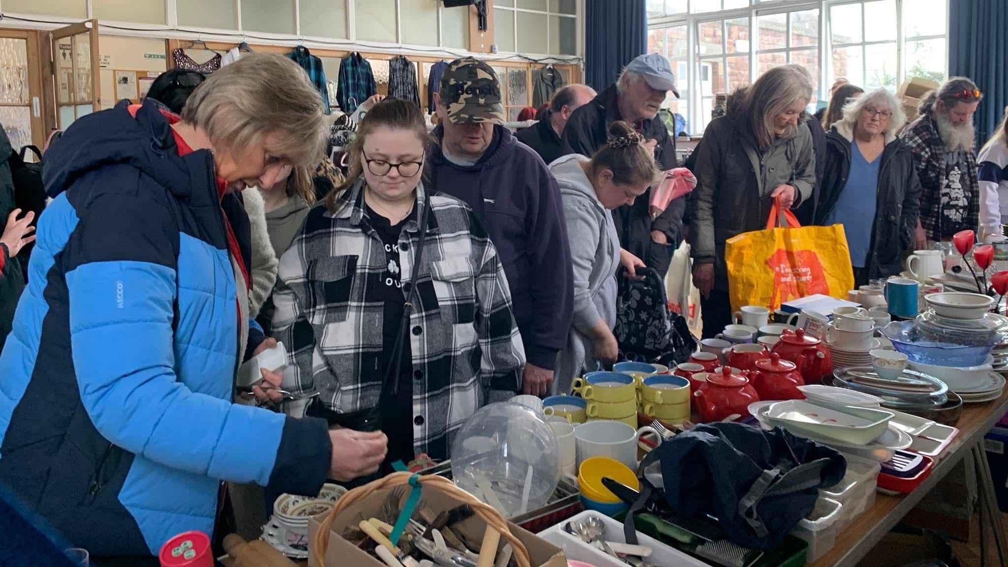A group of people looking at items on a table. Things such as teapots, hangers and other various household items are laid out.