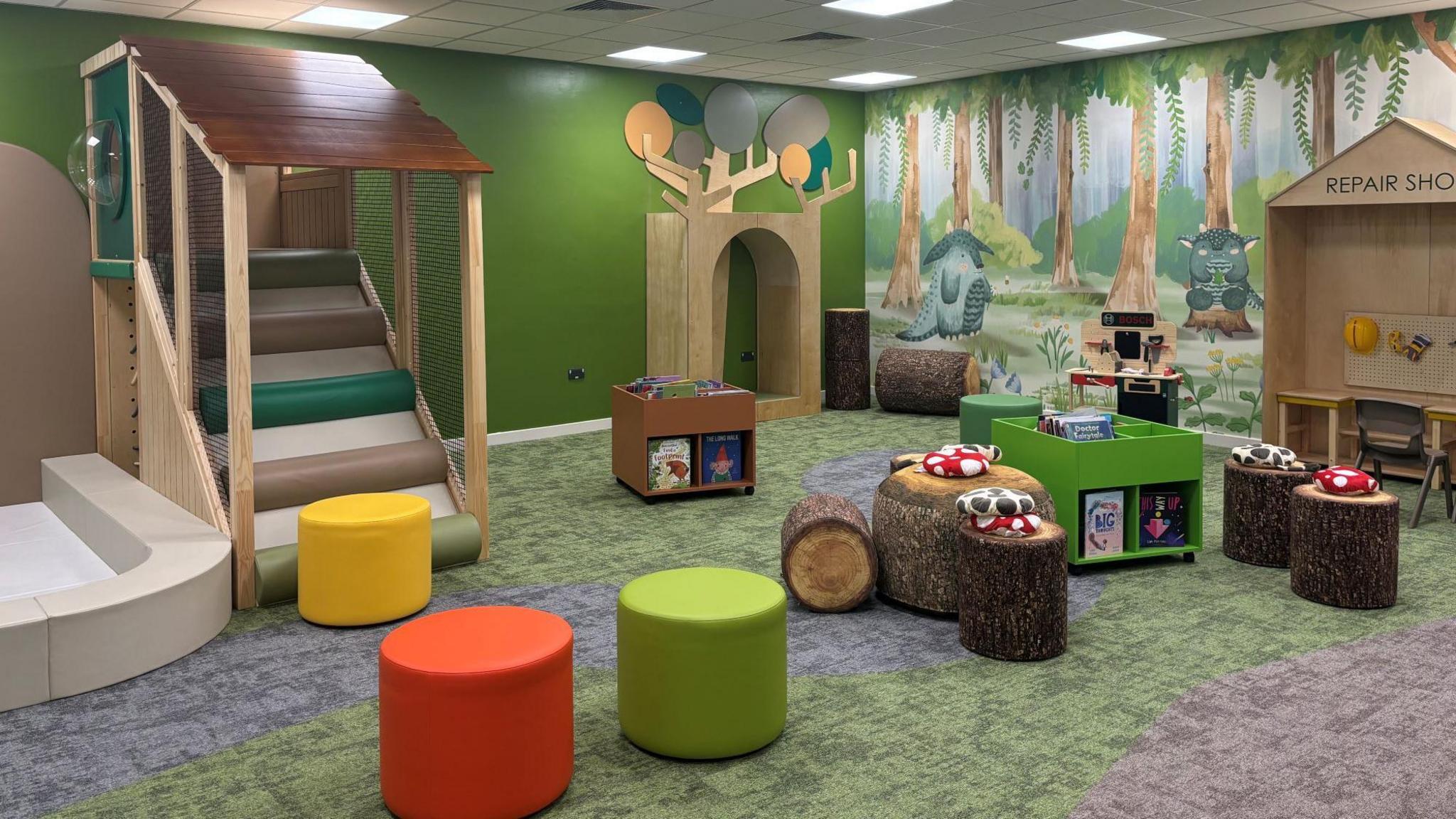 A children's area with multi-coloured stools and trees painted on the walls. There is a small staircase and stools that look like toadstools for the children to sit on. There are small shelves for books in the middle of the rooms. The carpet is mostly green.