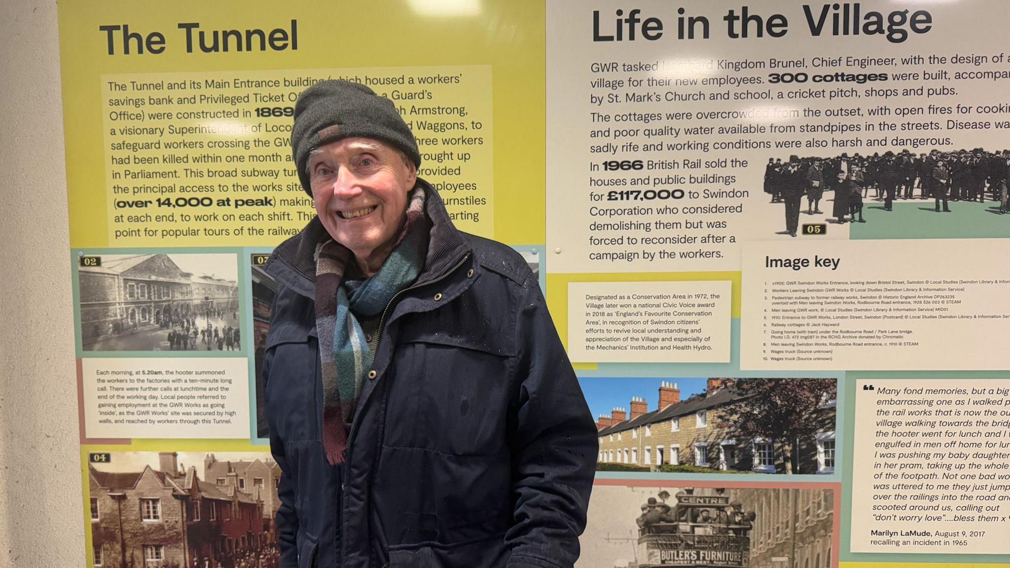 An elderly man in a dark coat, scarf and grey hat smiles at the camera. He is standing in front of green and white information boards that display historical pictures of houses, and text outlining Swindon history.