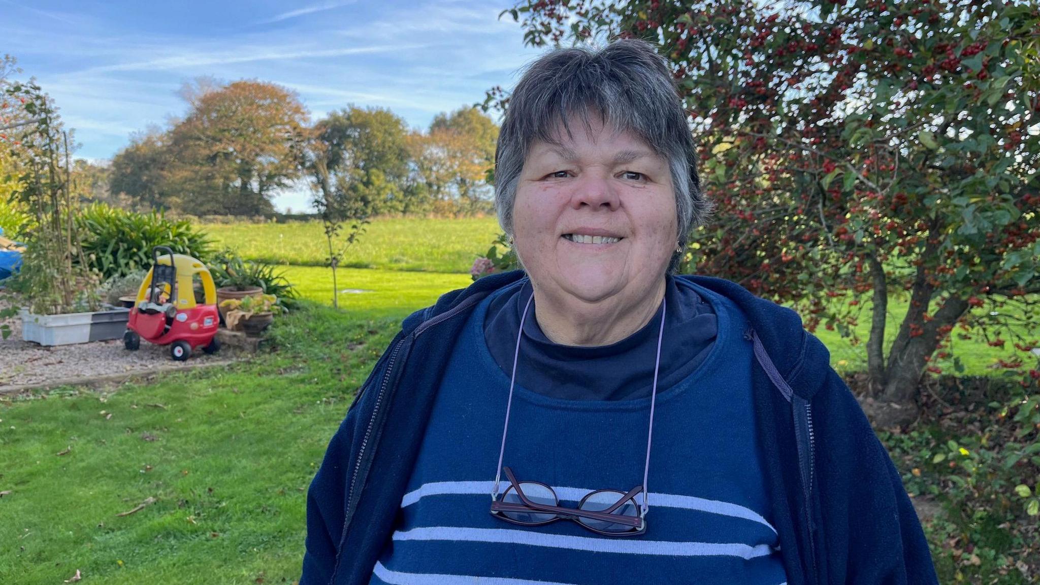 Monique Lawrence stands in a garden. She has short grey hair and is wearing a blue jacket over a blue jumper. She has a pair of spectacles on a lanyard around her neck.