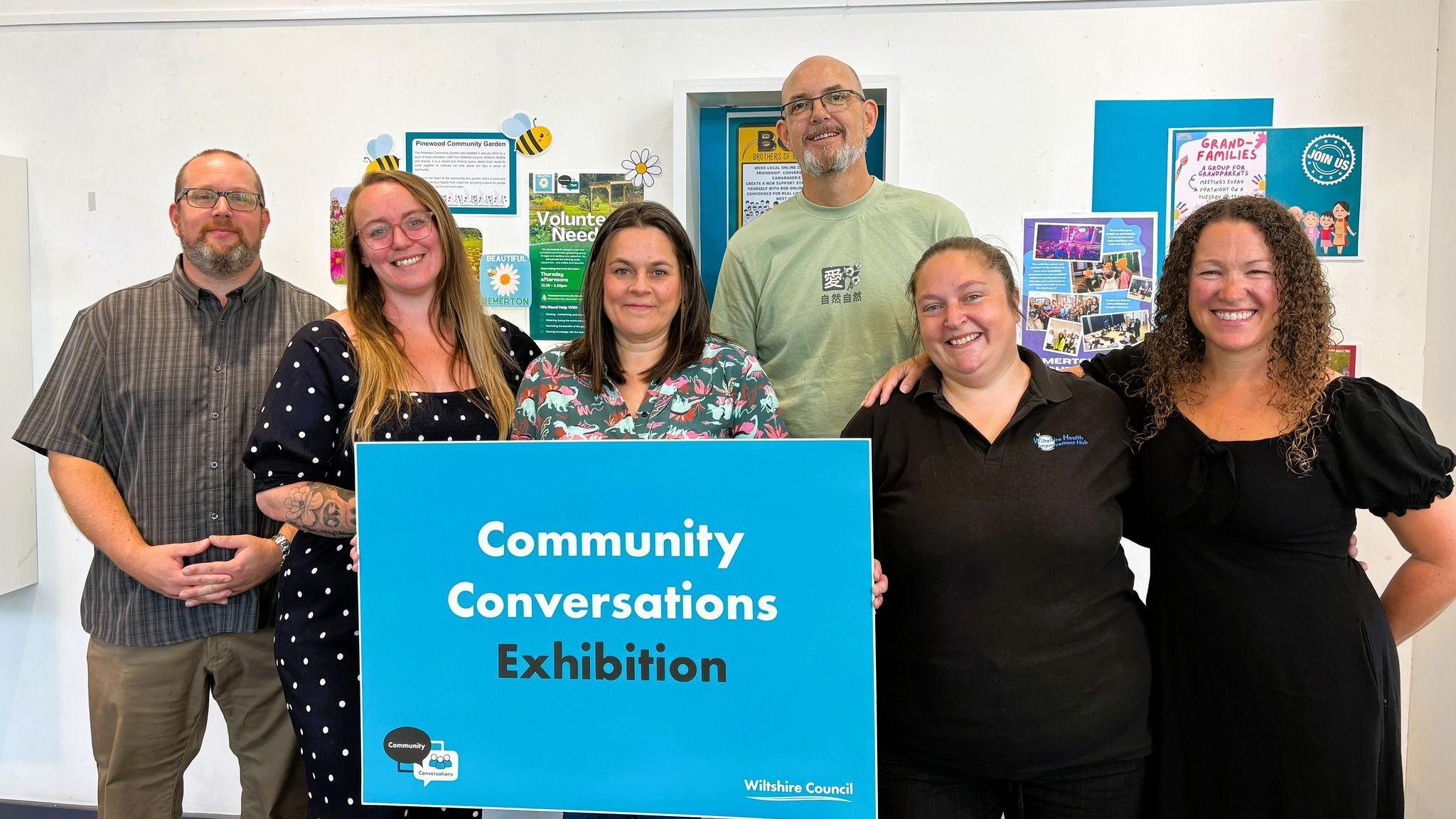 A group of people - four women, two men - stand together, with some exhibition posters on a white wall behind, in the middle, they hold a big blue sign saying 'Community Conversations Exhibition'