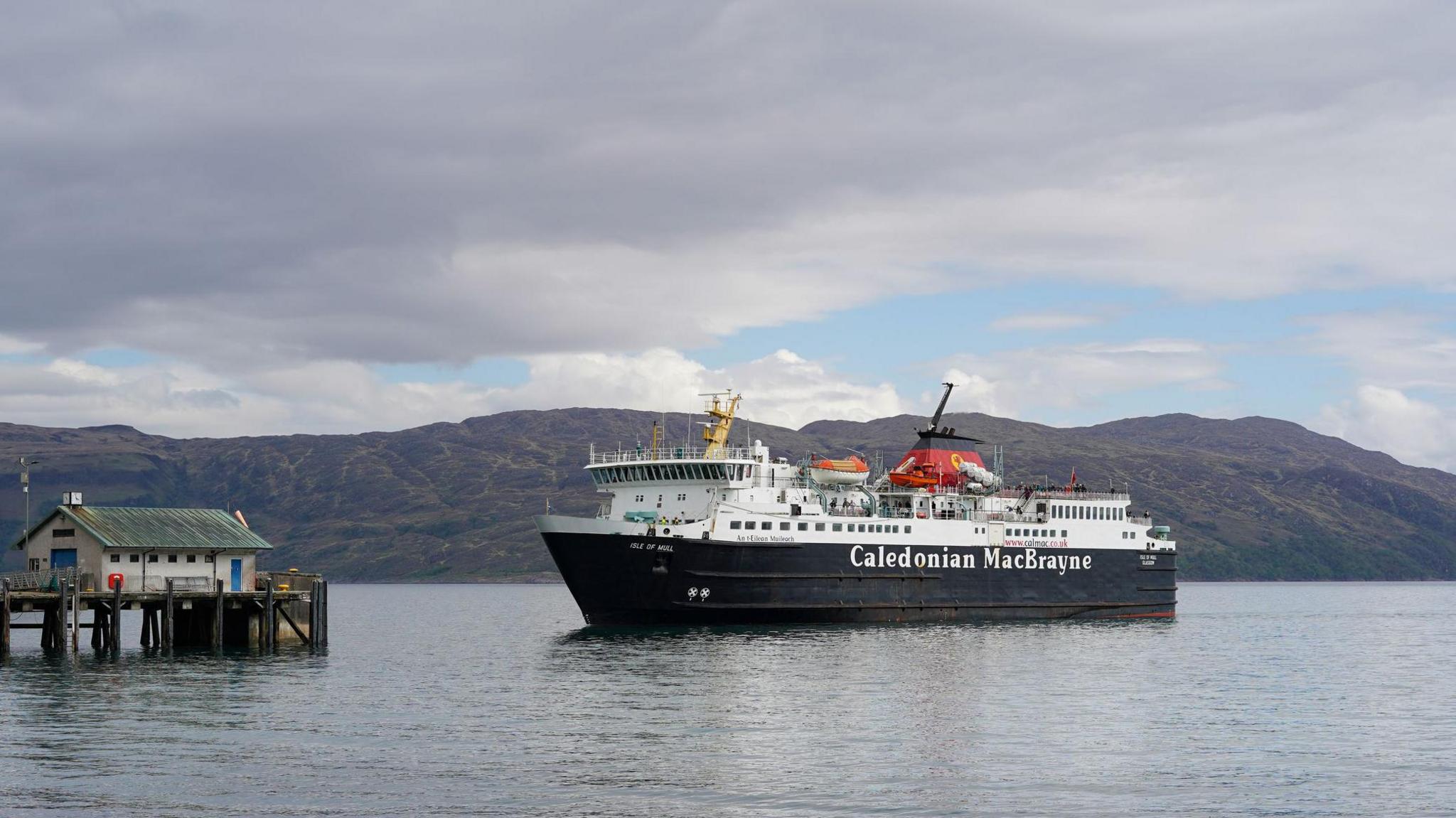 An MV Isle of Mull, bàta-aiseig ChalMac a' dol a-steach gu cidhe Chreag an Iubhair. Bàta dubh is geal le Caledonian MacBrayne sgrìobhte air a cliathaich. Tha bothan beag le doras gorm aig deireadh a' chidhe agus tha beanntan fada às