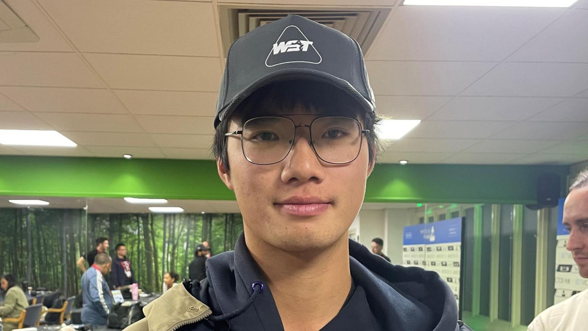 Xianghao Xiao wears a beige jacket over a black hoodie, and has a black cap on his head. He is standing in a media room at the Brentwood Centre. The room has green walls. Various people are sitting or standing in the background.