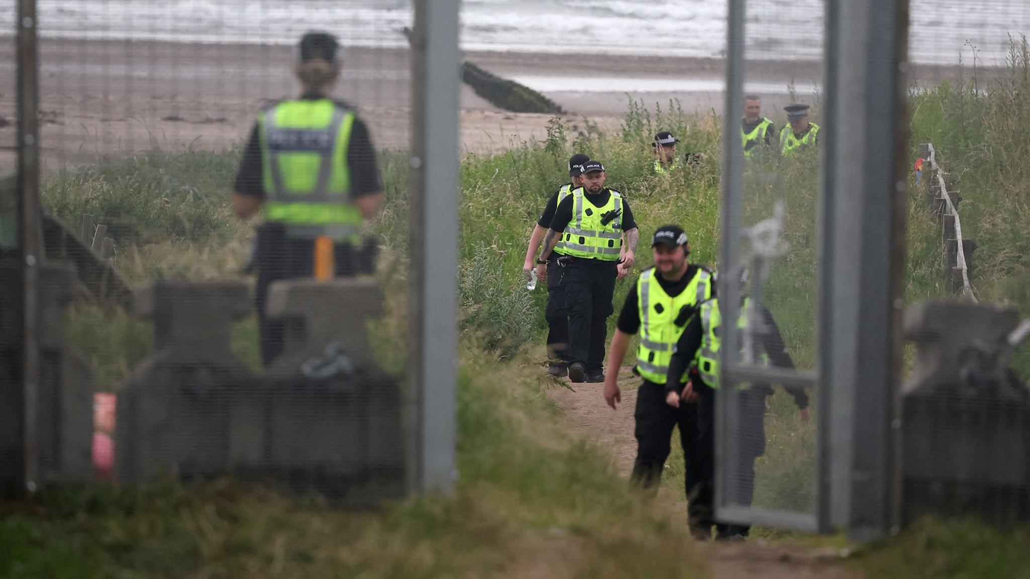 Police in high viz jackets on a beach behind a large fence