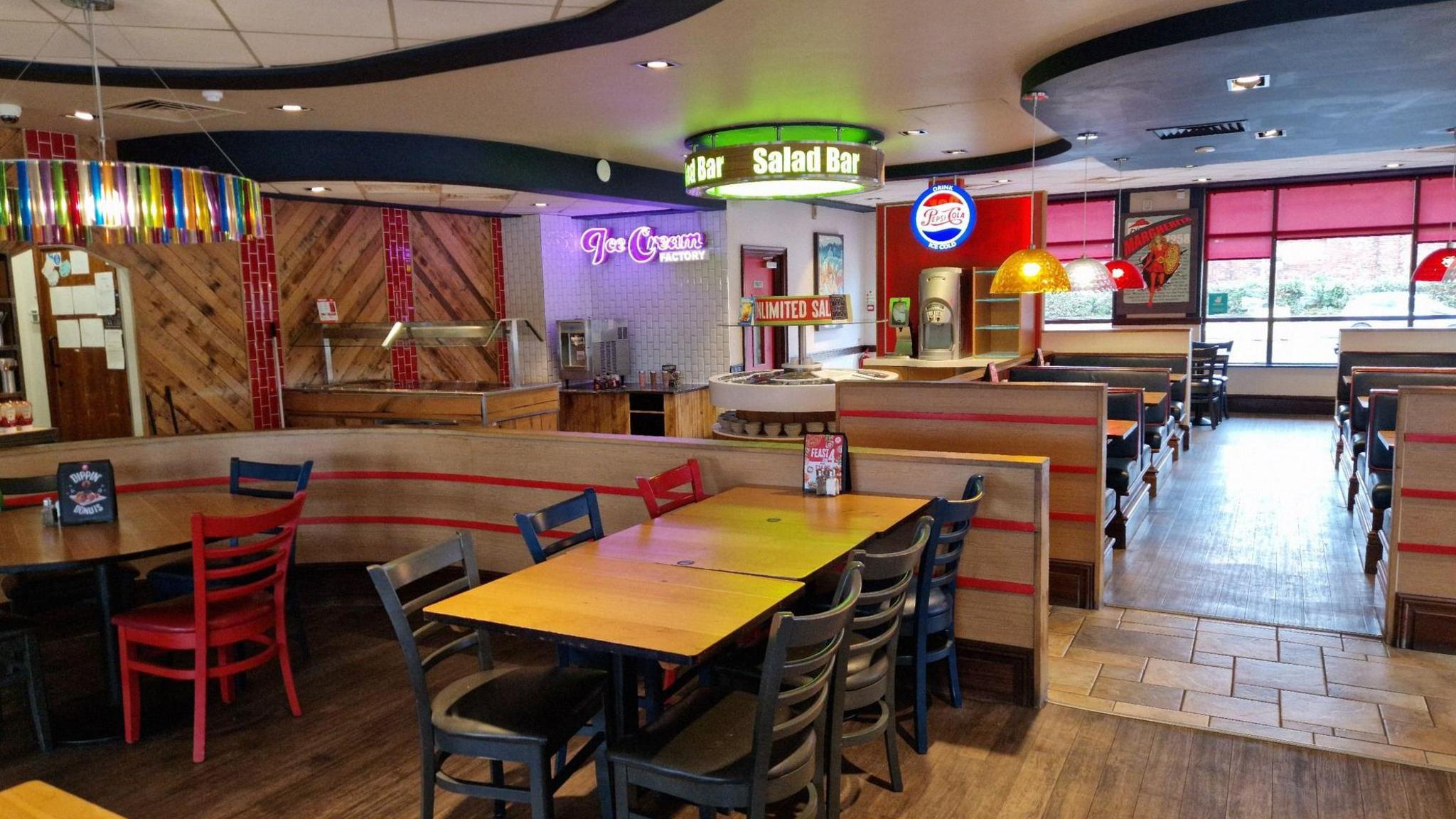 The interior of Hartlepool Pizza Hut. There are signs saying Ice cream Factory and Salad Bar on the roof. Blue and red tables and chairs are clean and ready for the day.