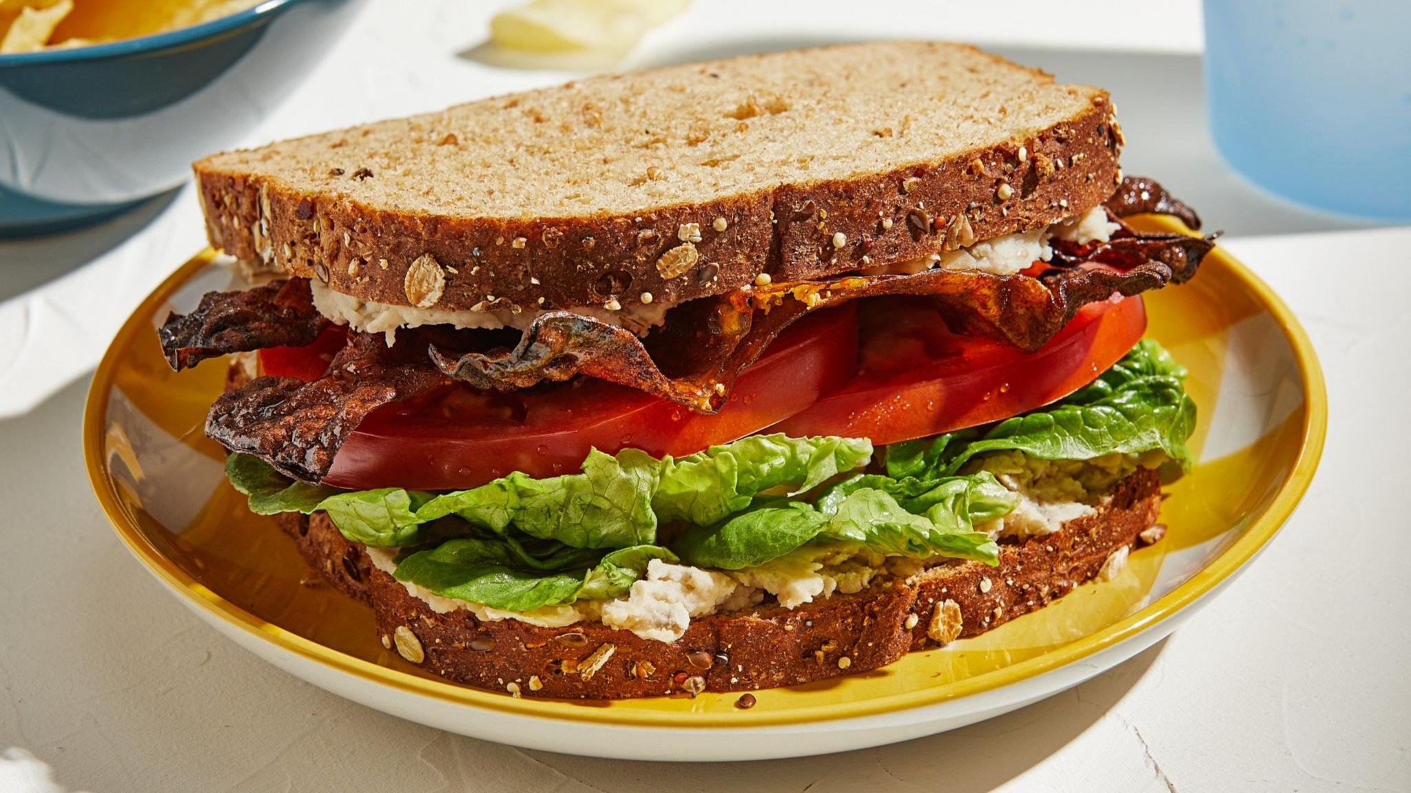 A bacon, lettuce and tomato sandwich on brown seeded bread. It is resting on a yellow plate on a table. 