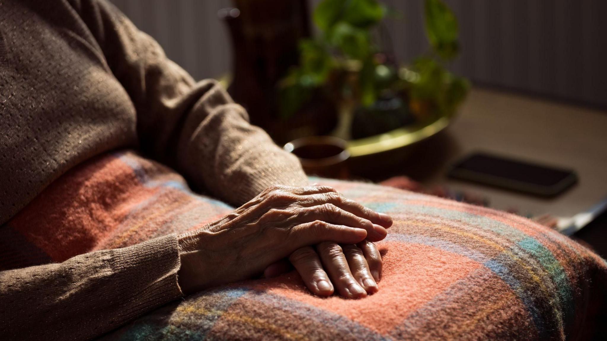 An elderly person sitting down with their hands crossed placed on a rug on their lap.