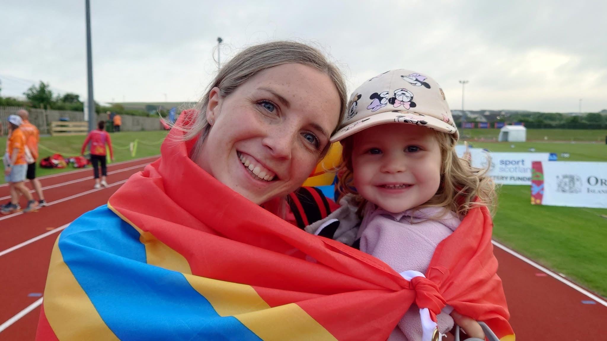 A woman holding a small girl, wrapped with a red, blue and yellow Orkney flag