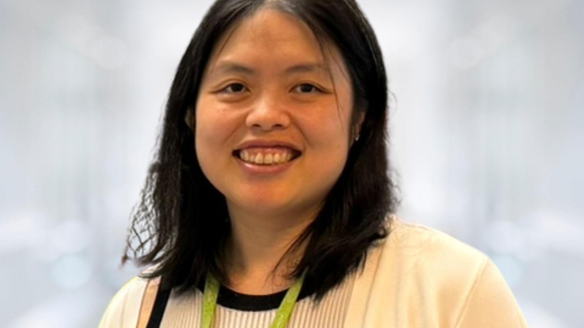 Dr Helena Lee has shoulder length black hair and is wearing a cream top, a cream cardigan and a green lanyard. She's smiling at the camera against a blurred white background.
