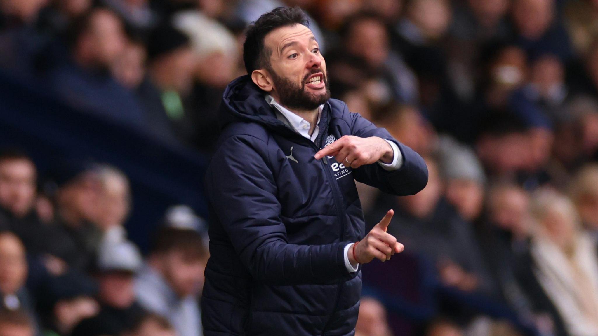 Carlos Corberan, wearing a blue West Brom coat, gives instructions to his team from the sidelines.