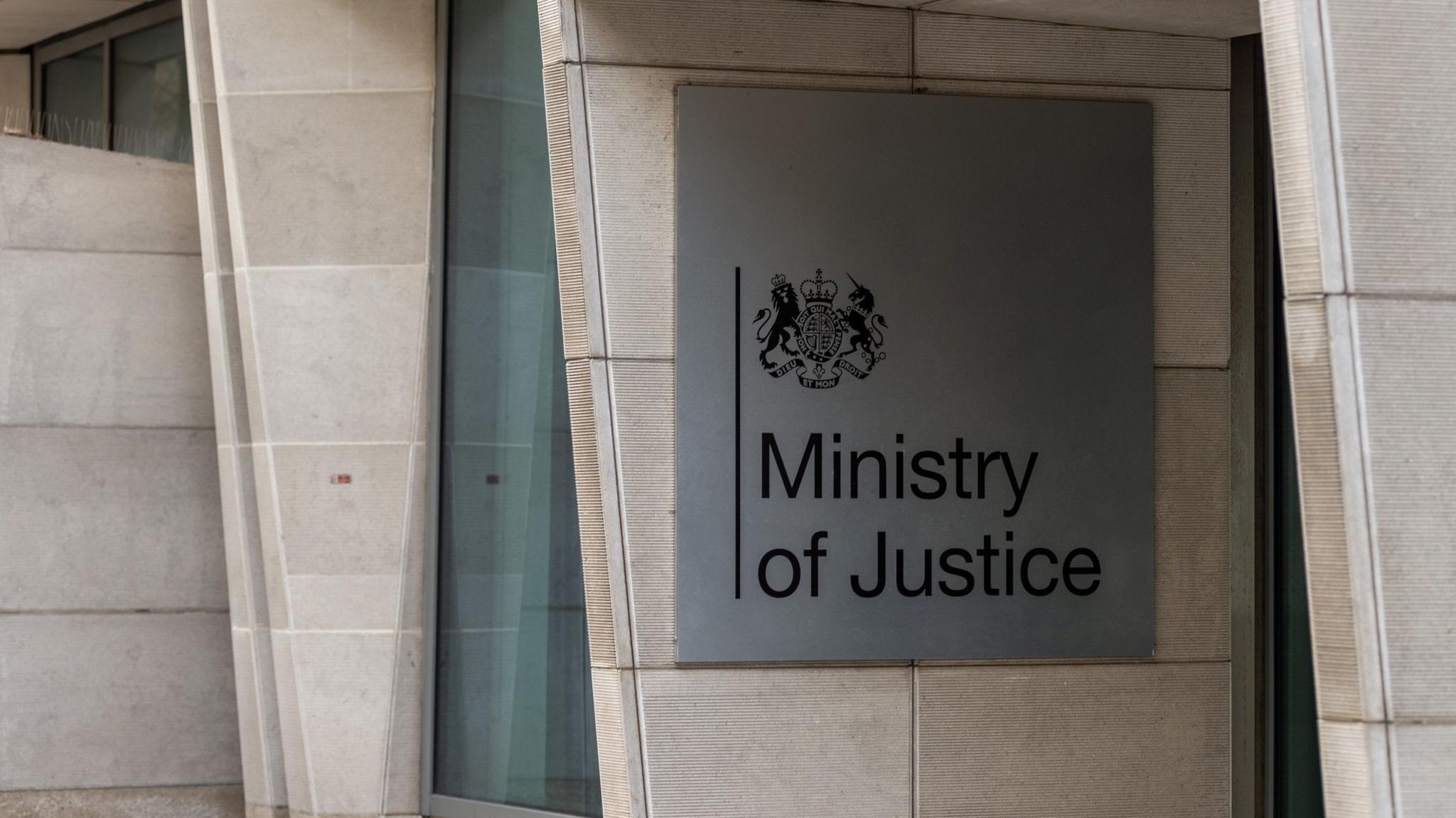 A square, silver-coloured sign with a royal crest reads "Ministry of Justice". It is attached to a light-coloured stone building.