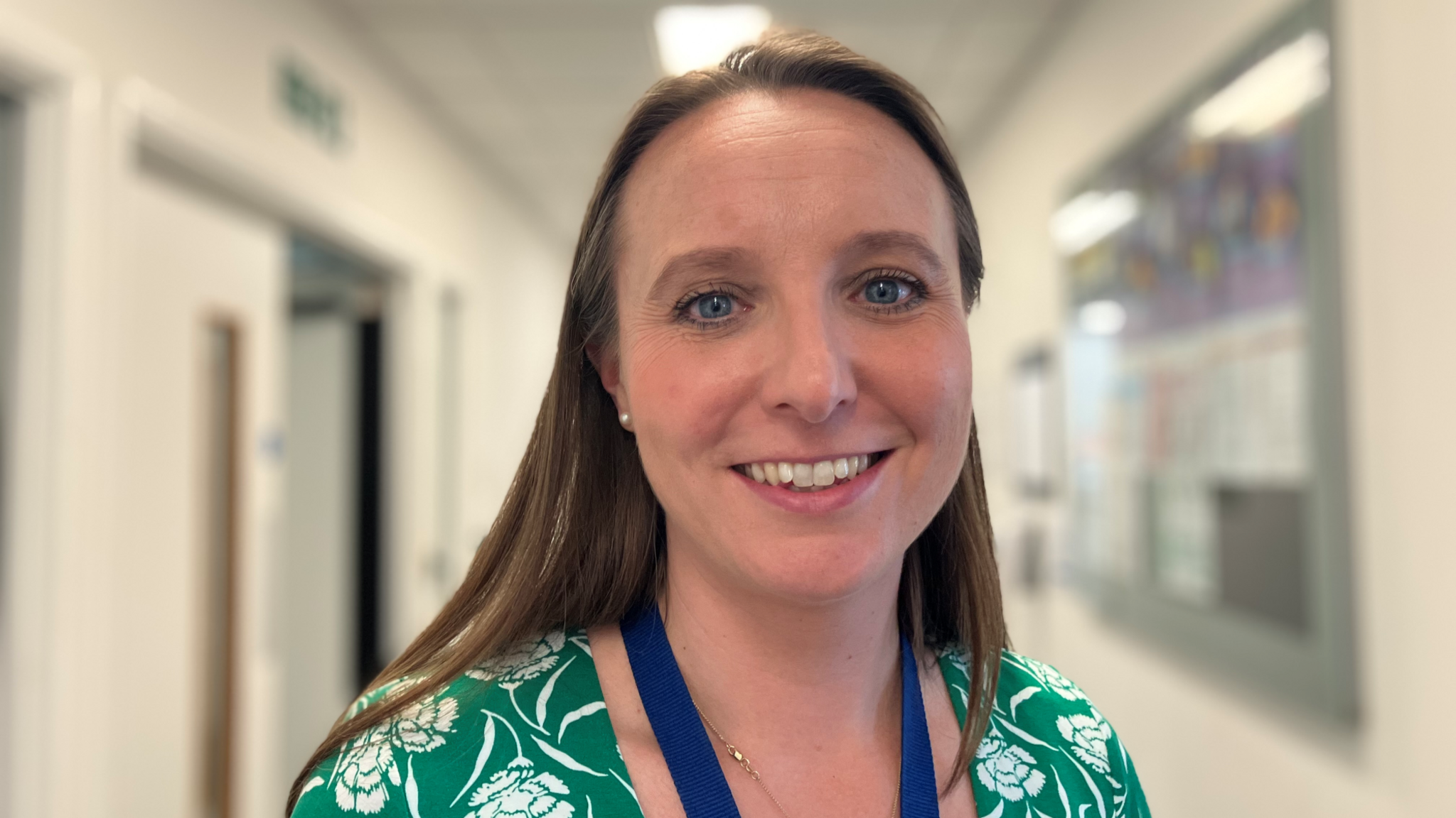 A woman with light brown hair wearing a green and white dress and a blue lanyard with a corridor in soft focus behind her