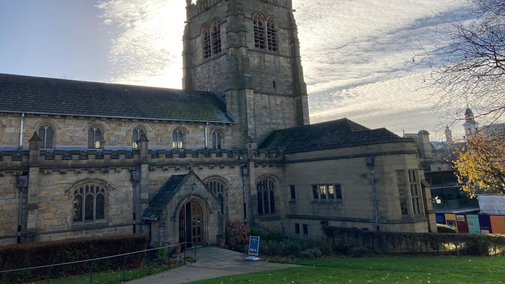 A stone-built building with a bell tower and a green lawn in front of it with a footpath leading up to an arched entrance. 