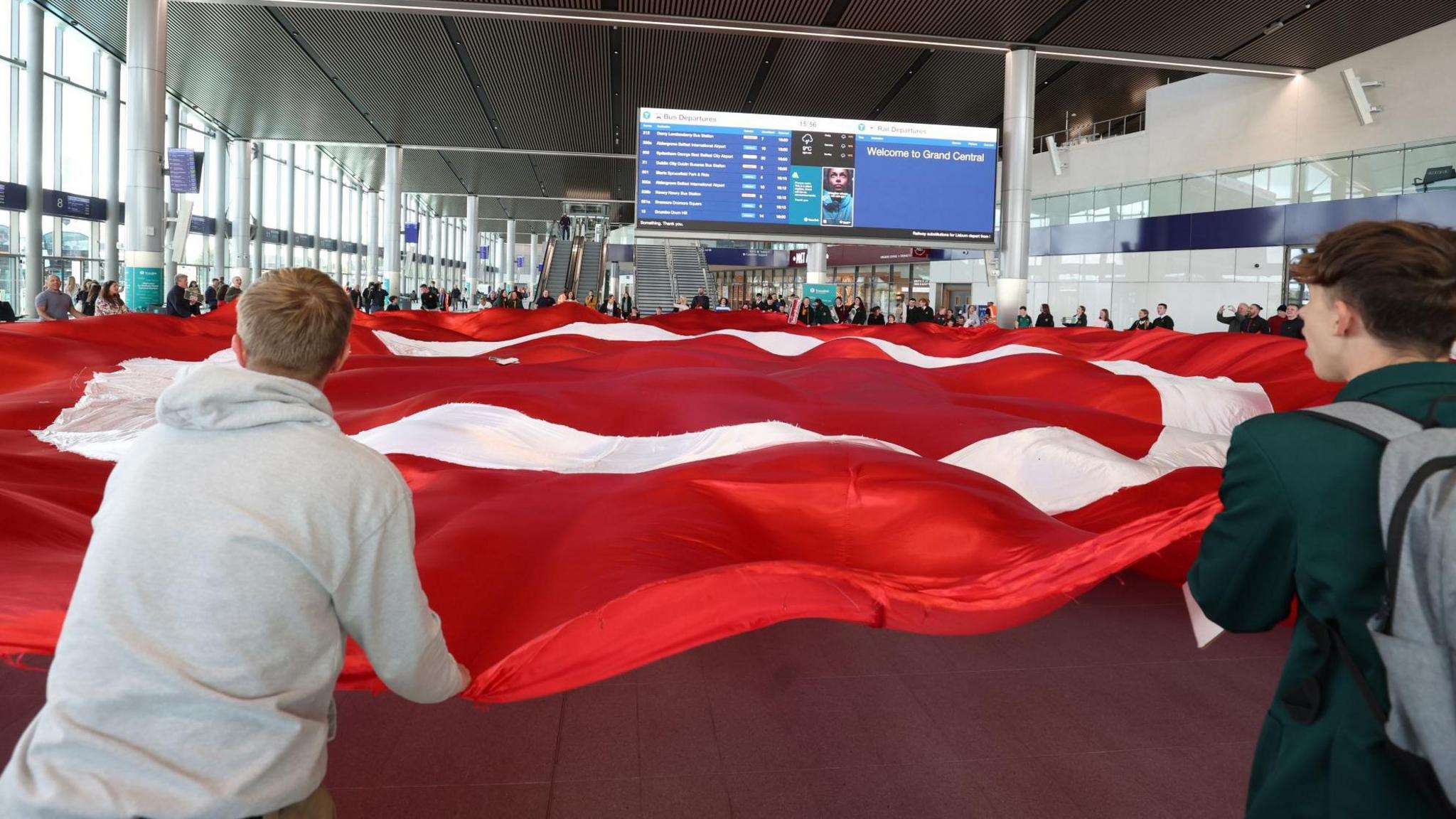 Protestors holding a large red flag with a white circle in the middle stand in the middle of the large main hall of Grand Central Station. The flag is the logo of Irish language group An Dream Dearg