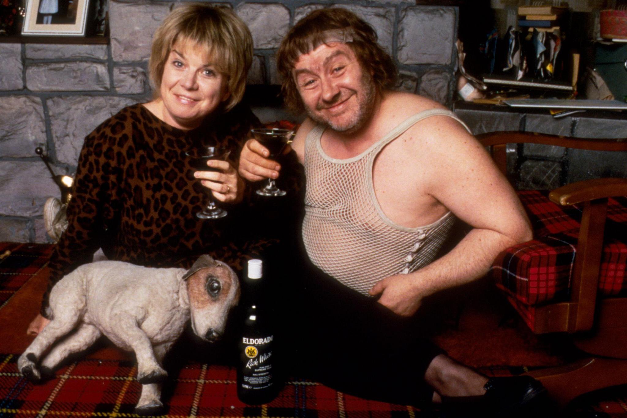 A woman with blonde bobbed hair raises a glass of Eldorado wine with a scruffy-looking man in a white string vest and grubby headband. They appear to be in a pub setting. An ornamental Jack Russell dog is lying down on a tartan tablecloth in front of them and next to the wine bottle. 