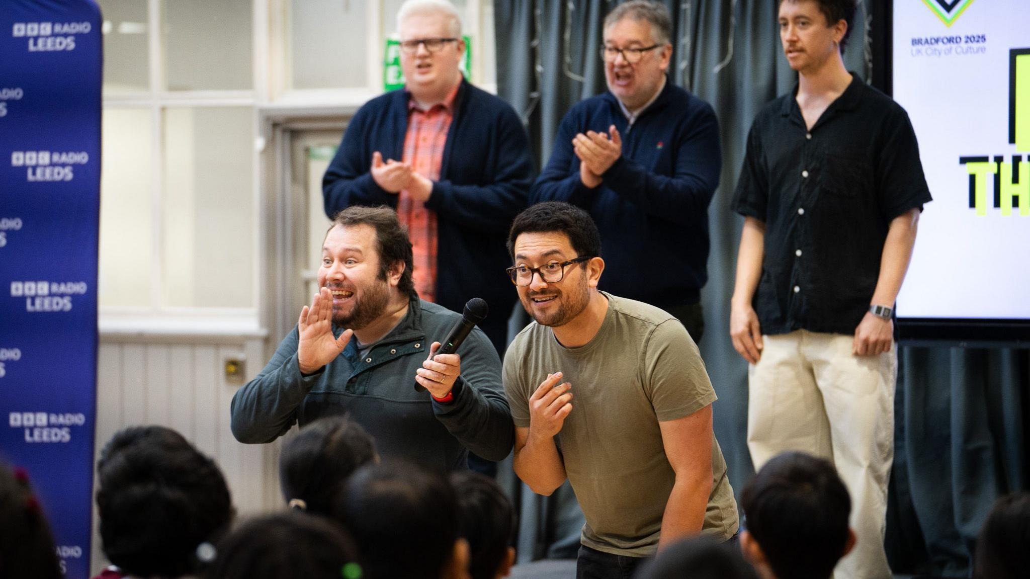 Three men stand on a stage clapping, with two men stood on the floor in front of them addressing a crowd of school pupils. One of the men is holding a microphone. In the background is a large screen with the Bardford 2025 City of Culture logo on it. A banner with BBC Radio Leeds' logo stands on the other side.