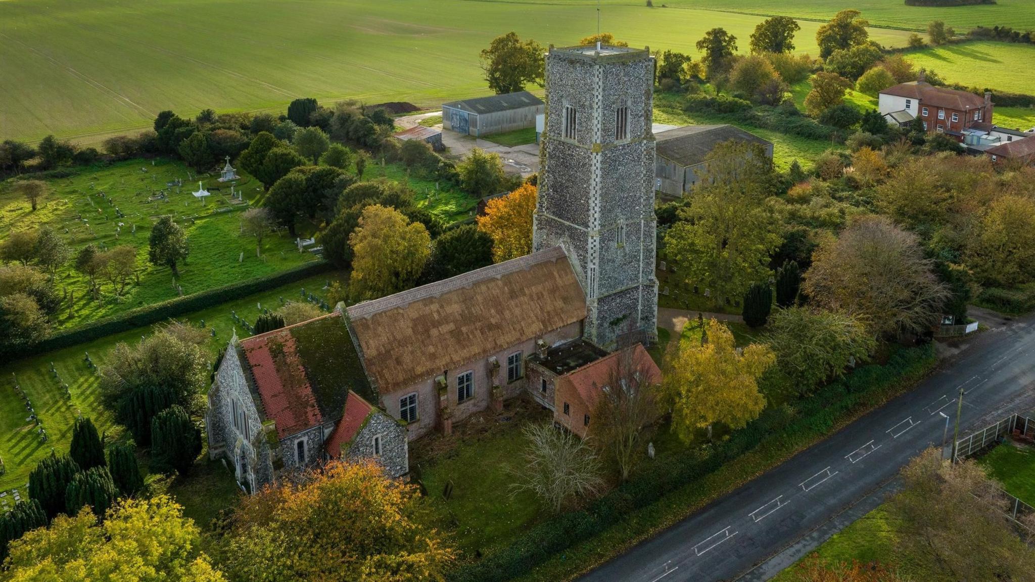 A aerial shot of St Edmund's Church, Kessingland, Suffolk. The church has a flint tower on the right and its partly newly-thatched roof stretches to the left. It has several colourful autumnal trees in front of it and behind. Beyond it can be seen fields and in the top right barns and a detached house