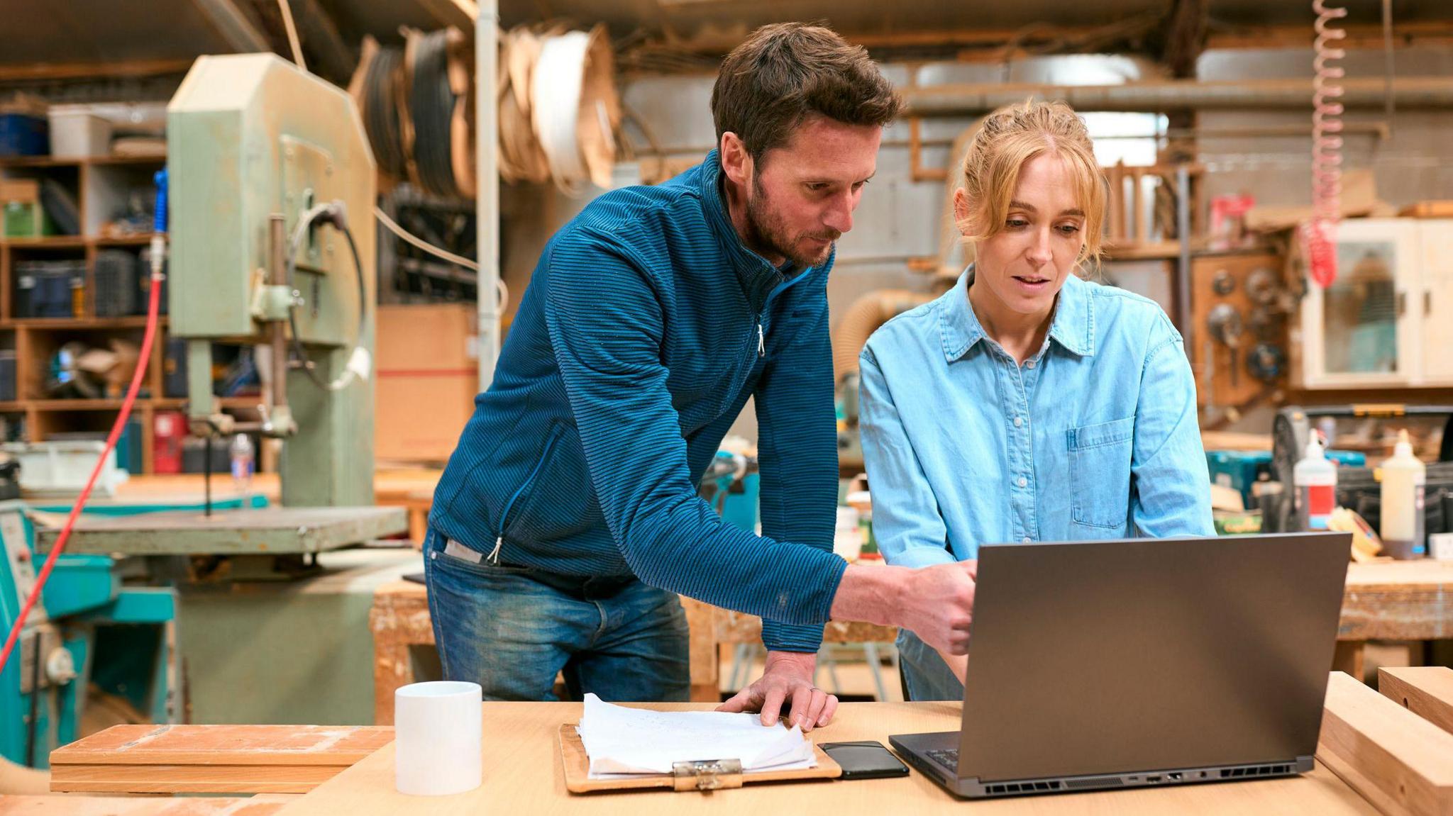 A man in a dark blue quarter zip long sleeve top showing a woman in a light blue denim shirt something on a laptop screen in a workshop with machinery behind them