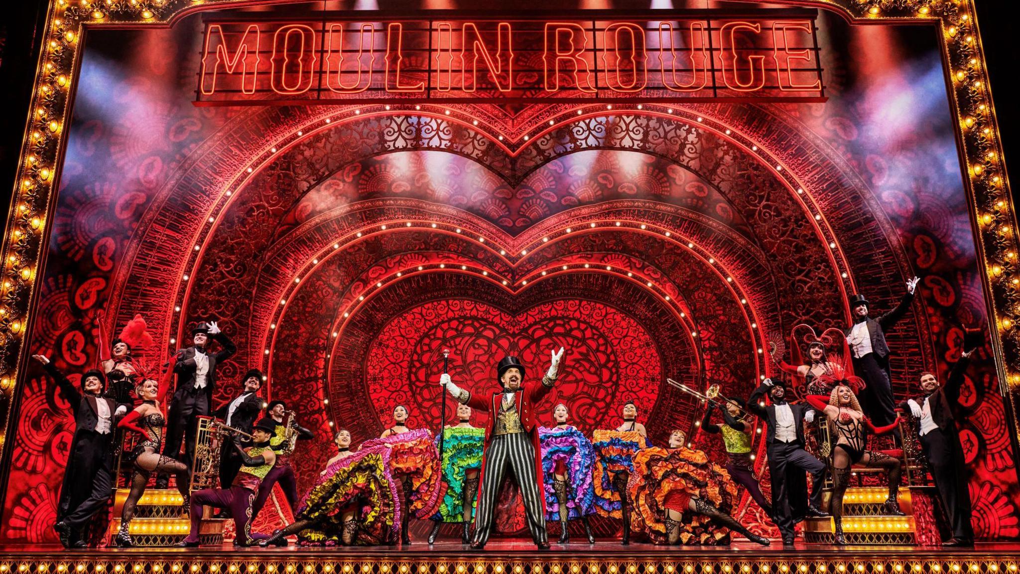 Performers in glitzy outfits stand on stage as they perform in Moulin Rouge at the Bristol Hippodrome. Behind and above them is an impressive set largely red in colour with "Moulin Rouge" spelt out at the top.