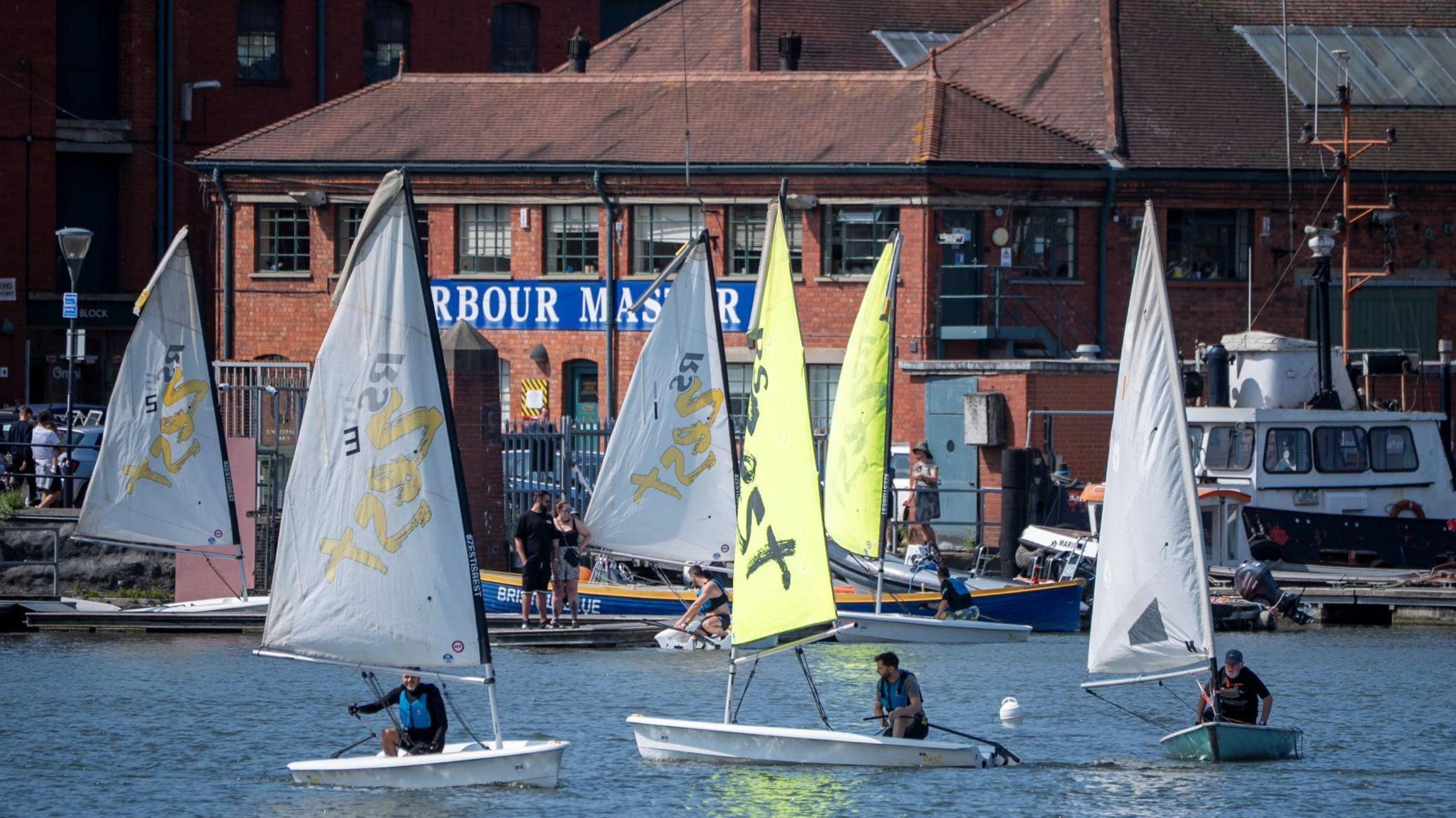 Sailing boats on the water in Bristol harbourside. The sails are a mix of yellow and white. In the background is the Harbour Master building. 