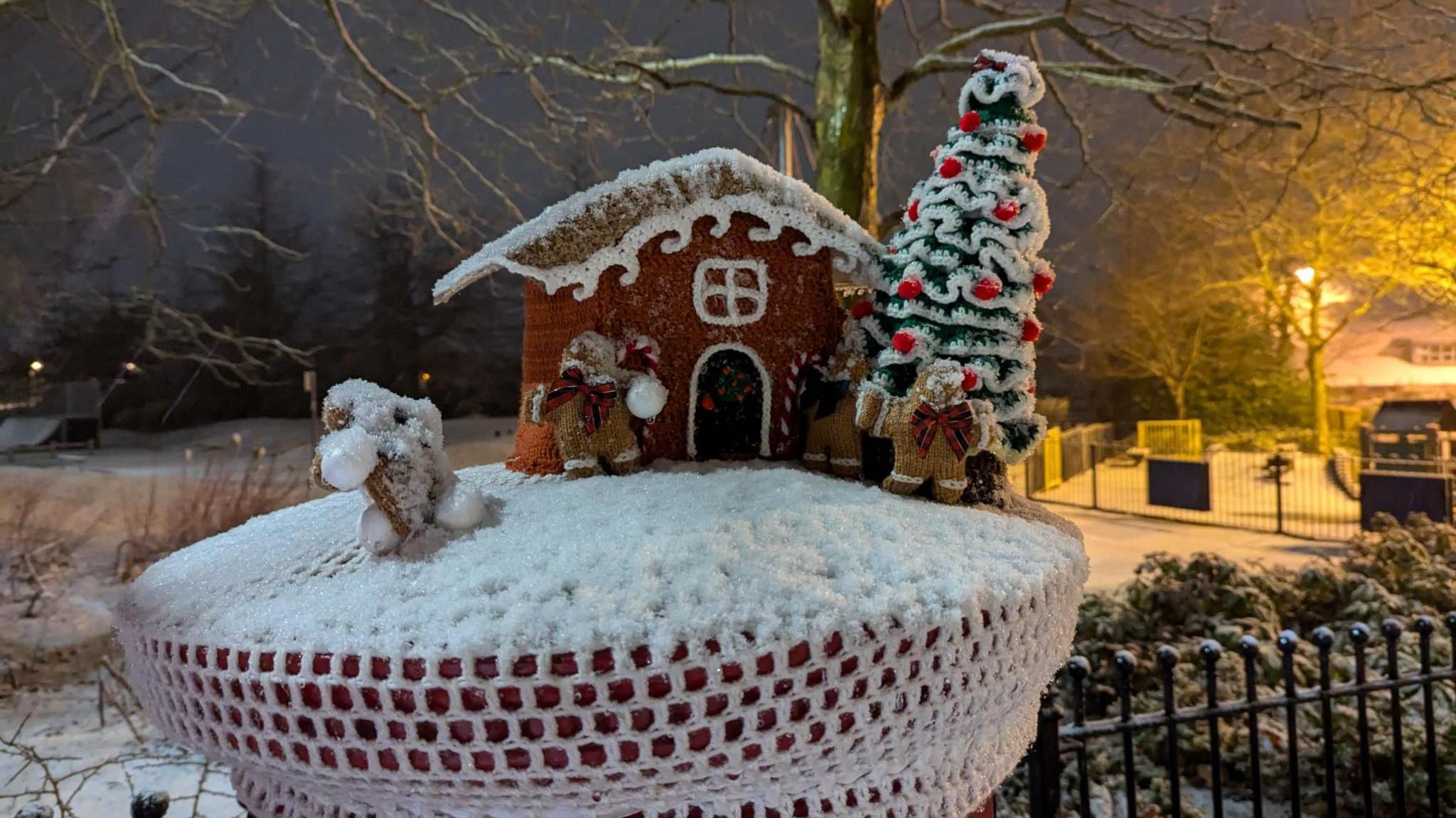 A photo of a festive postbox topper, featuring a Christmas tree and a gingerbread house, with a light dusting of snow on top. There's a snowy field behind it.