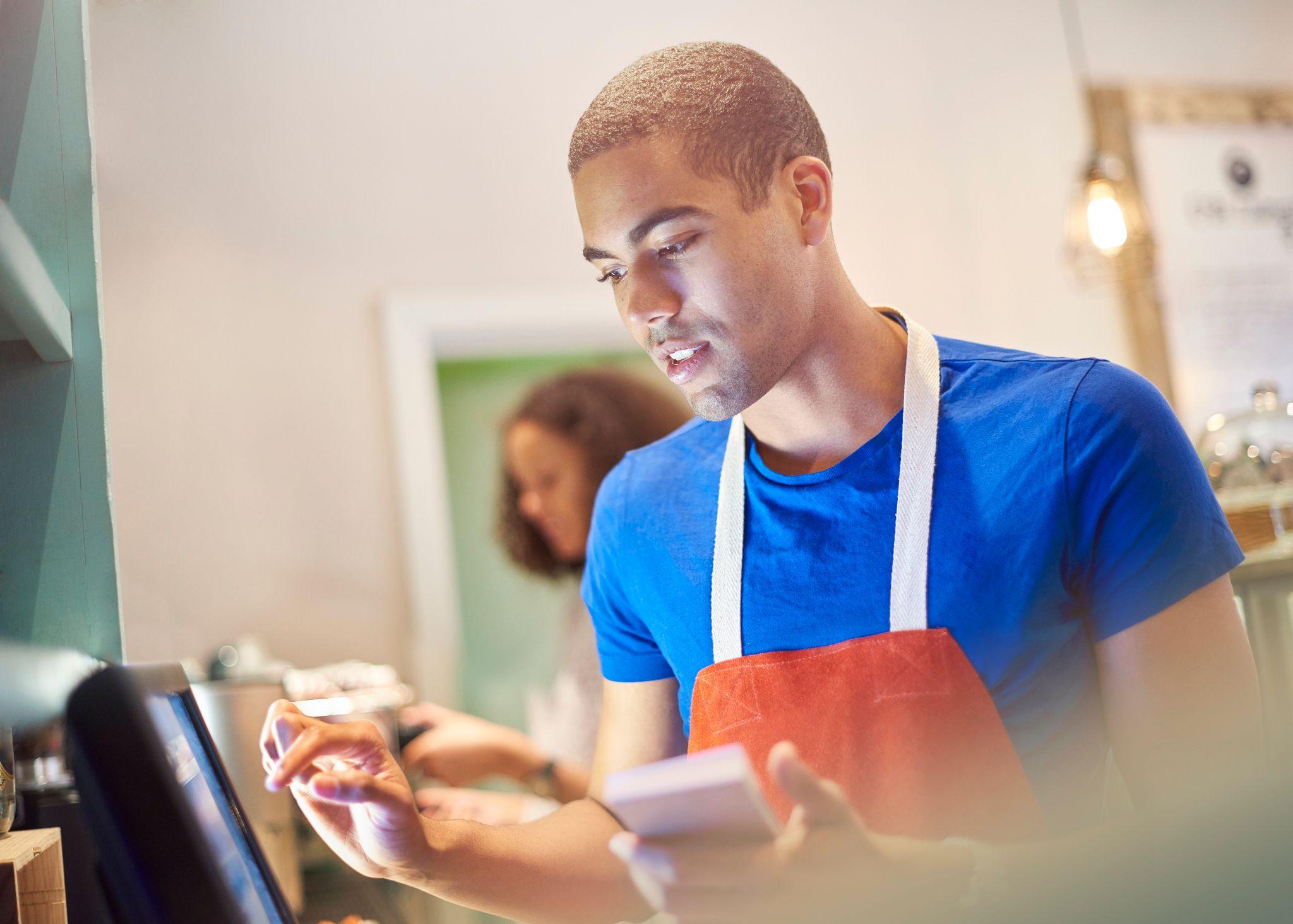 Young male waiting staff wearing a blue T-shirt and red apron, holding a notepad in one hand and typing on a touchscreen with the other hand