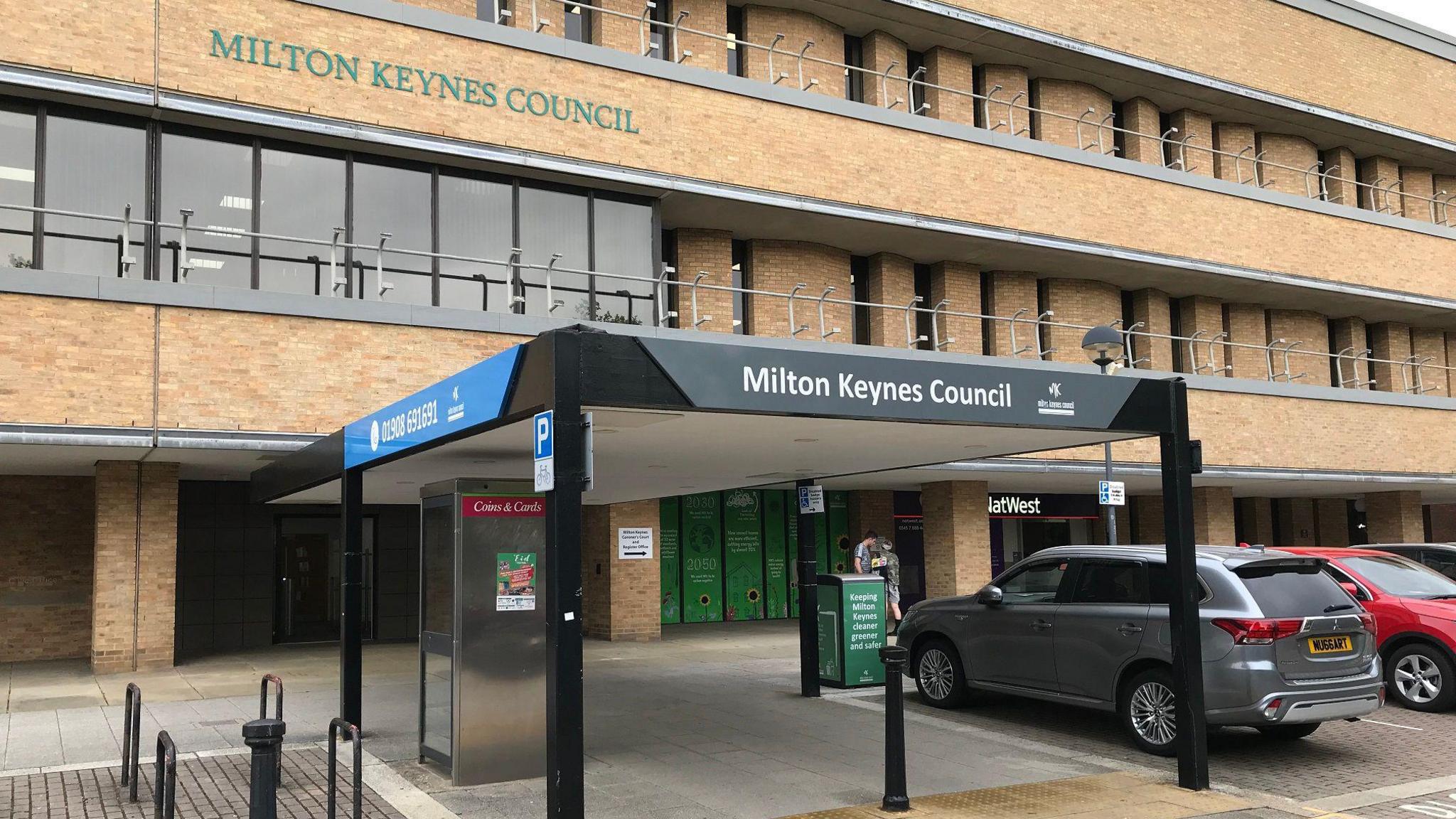 A picture of the outside of Milton Keynes City Council headquarters. The building is several stories high, with light brown bricks, with a covered entrance outside and a car park containung two vehicles.