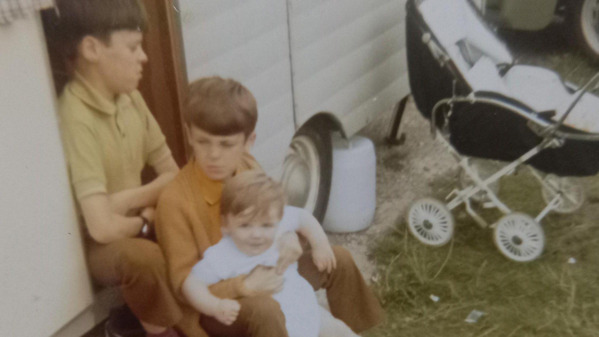 A vintage photo of three young boys sitting in front of one another on steps leading into what looks like a caravan.