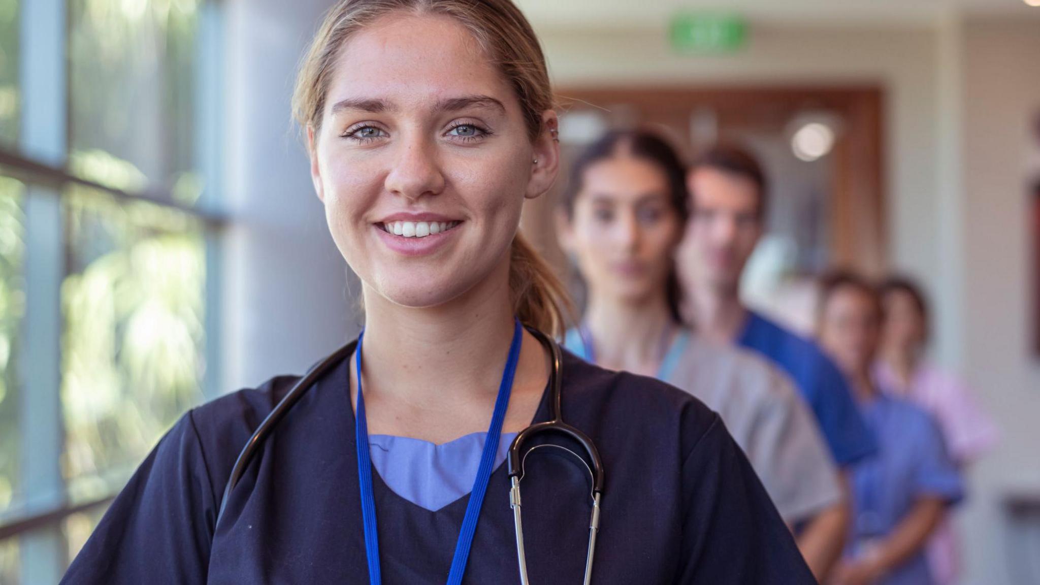 Health professionals standing in a line in a hospital corridor, with a woman at the front smiling at the camera.