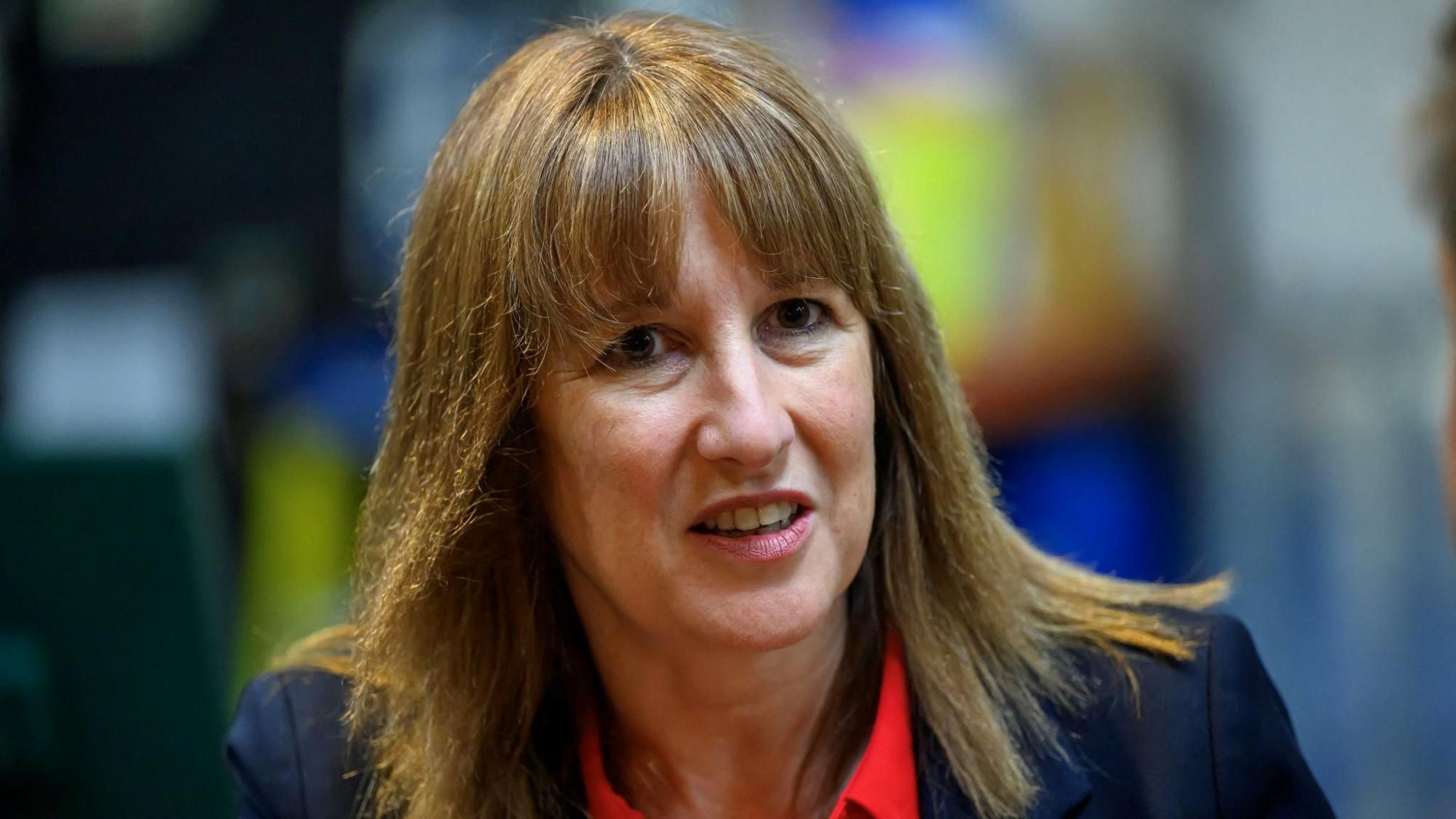 Rachel Reeves looking at the camera. She has shoulder-length brown hair with a fringe, and is wearing a dark blue jacket and a red top.