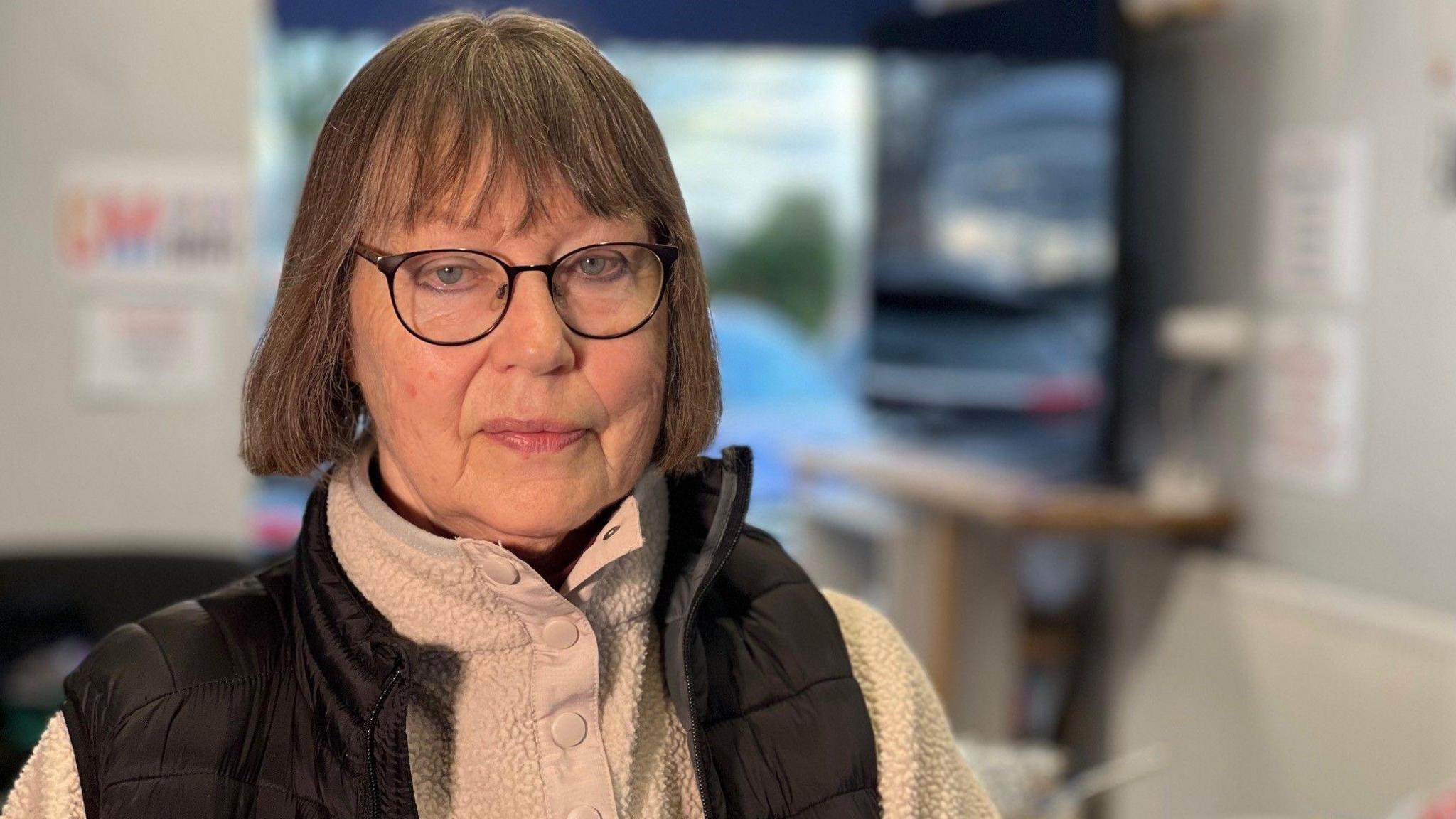 Annabel Marsh has bobbed brown hair and is wearing glasses. She is wearing a cream fleece top and a black padded gilet and standing in a room. A TV screen can be seen blurred out behind her.