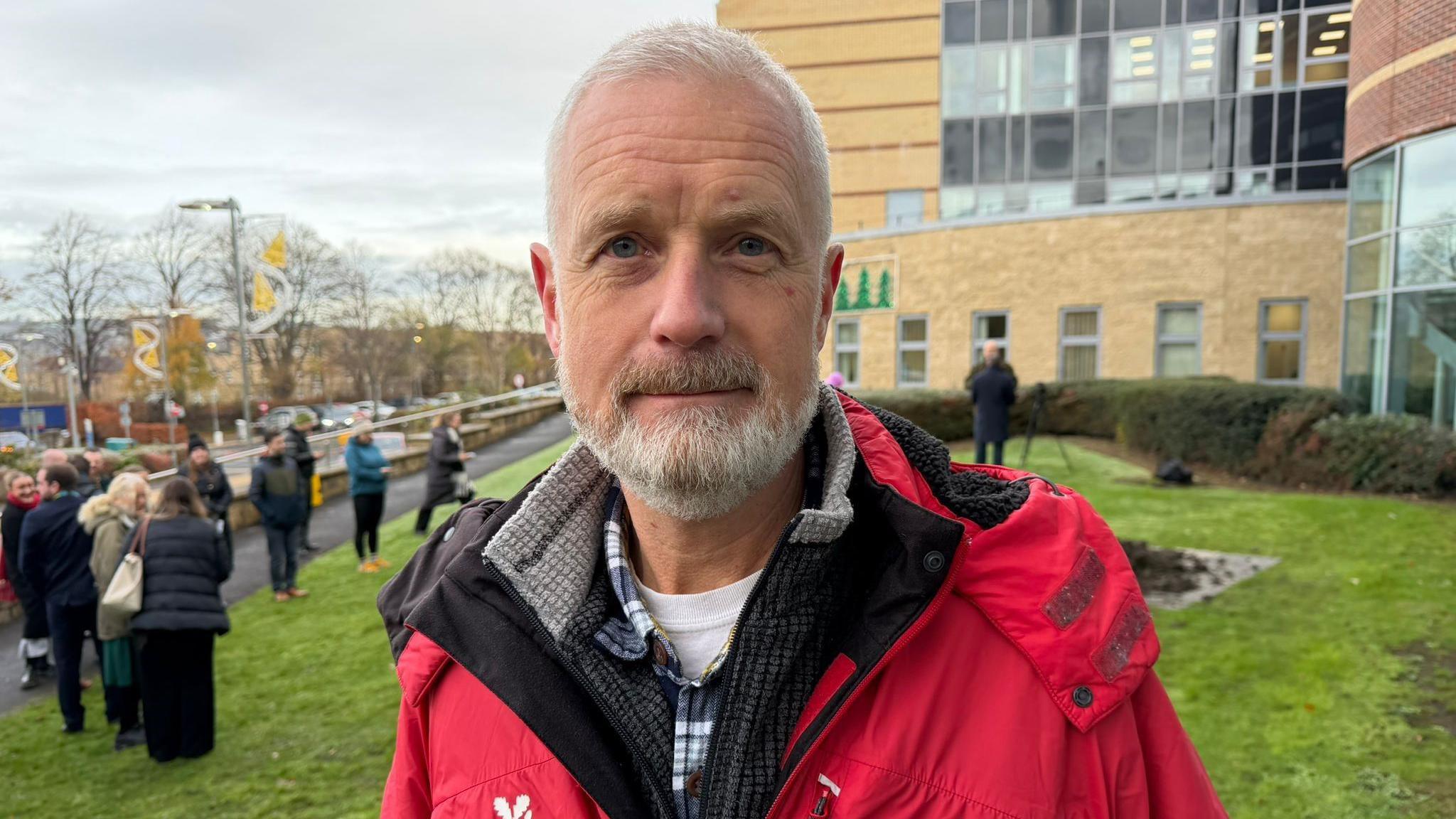 Andrew Poad looking into the camera. He has white, short hair and a white beard. He is wearing a red coat with a checked blue shirt visible underneath. Behind him there is a small crowd of people.